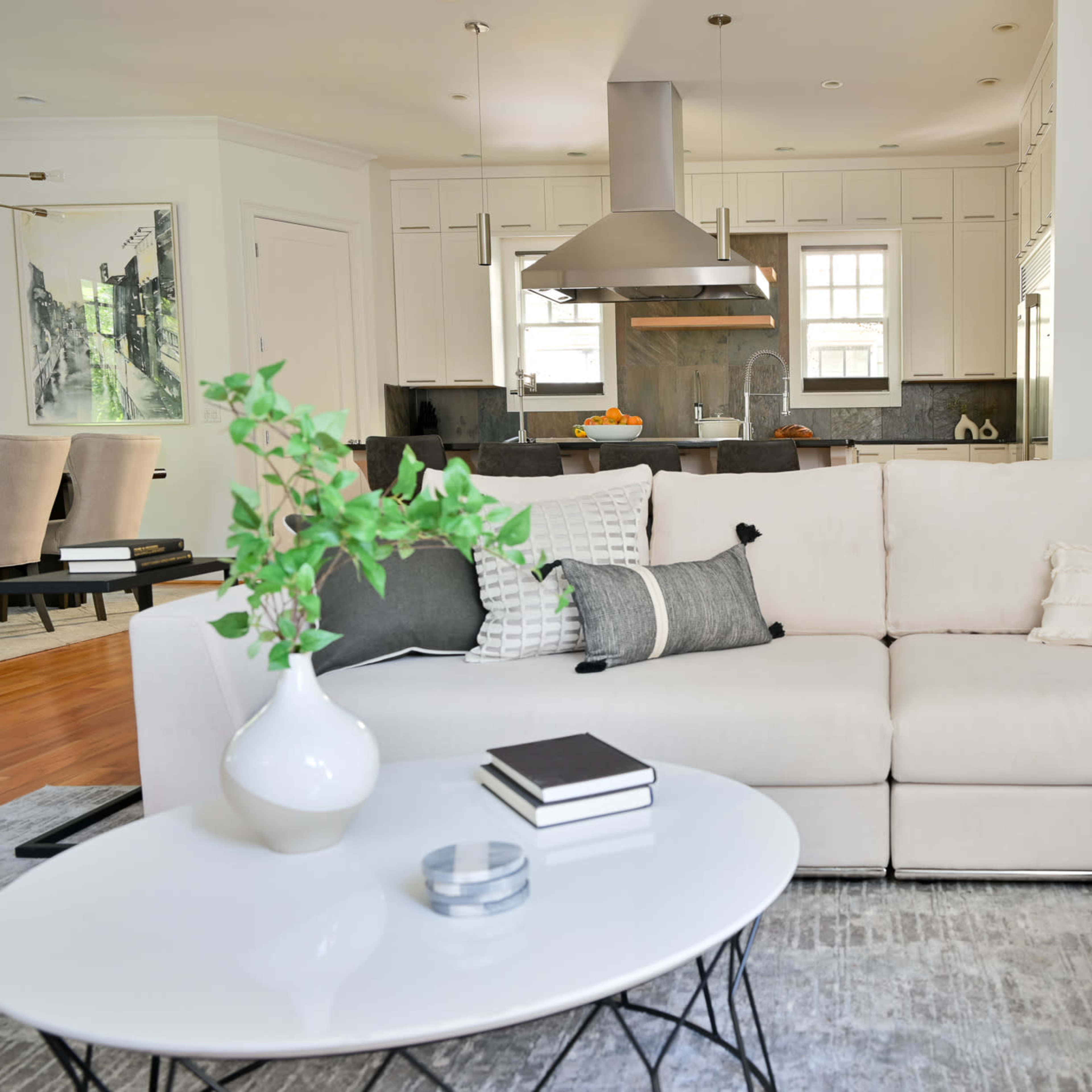 A modern living room features a light-colored sectional sofa, a round coffee table with books, and a view of a kitchen in the background.