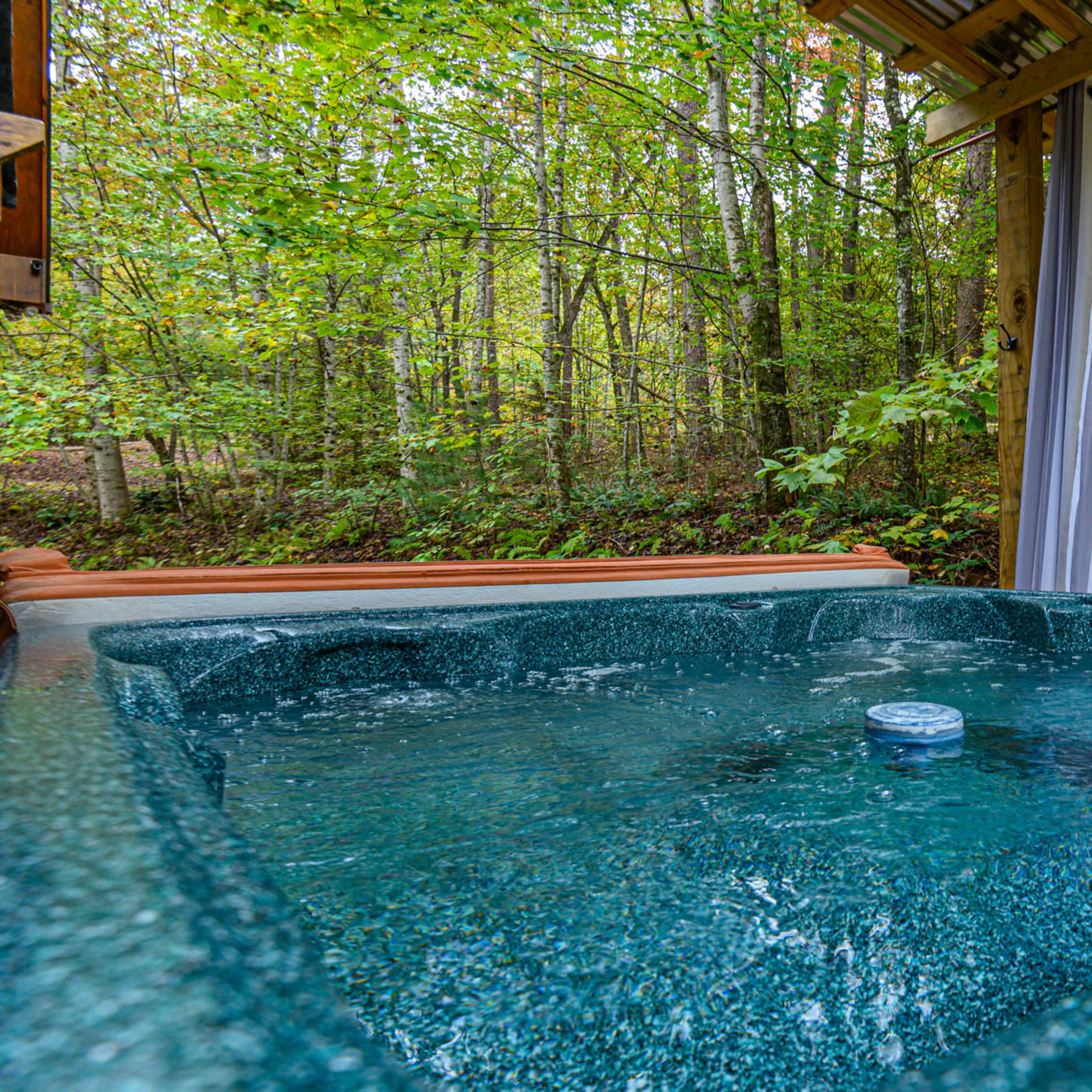 A hot tub sits under a wooden shelter surrounded by trees and green foliage.