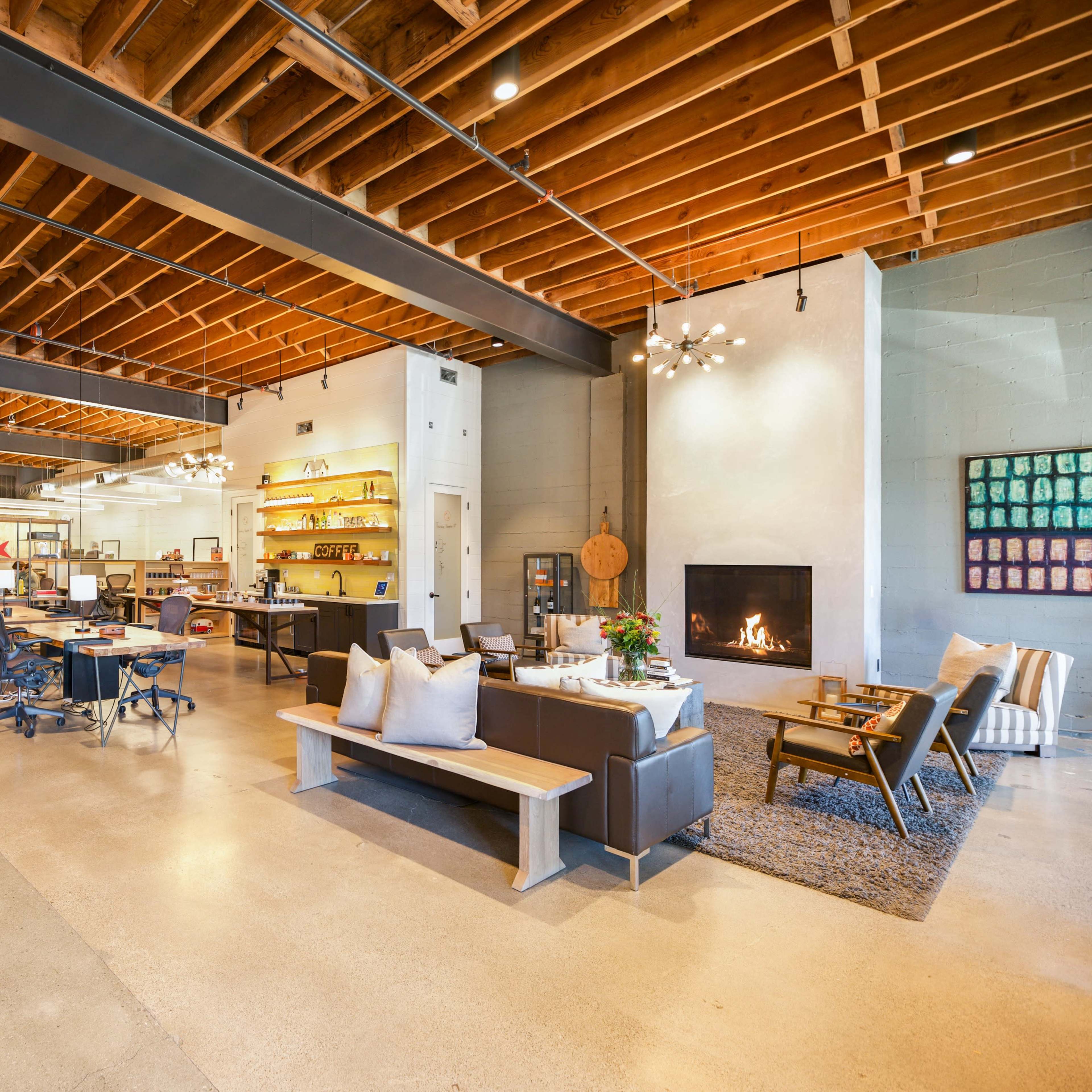 A modern, open-concept workspace featuring seating areas, a fireplace, and a kitchen area with industrial design elements and wooden beams overhead.
