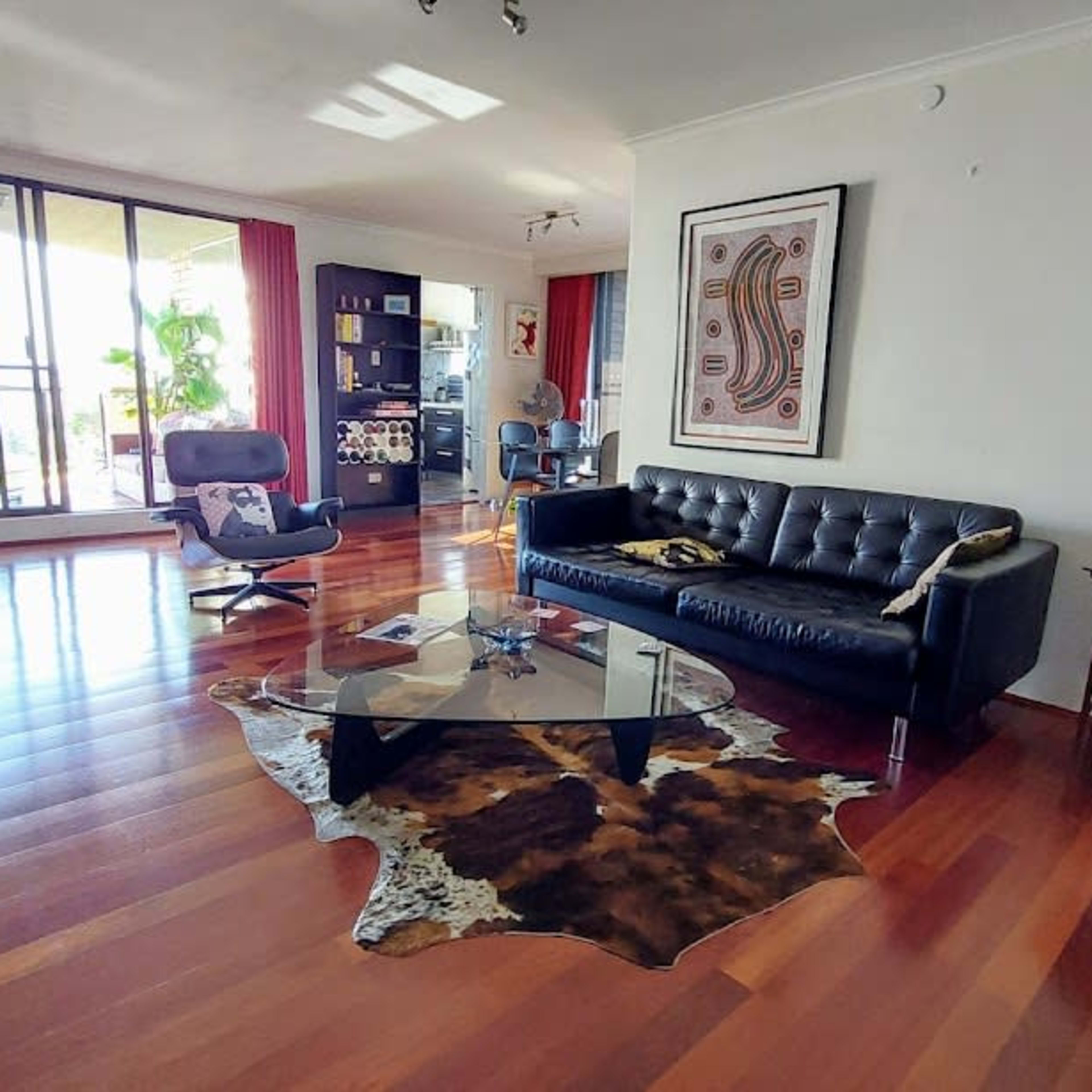 A modern living room featuring a black leather sofa, a glass coffee table, wooden flooring, and large windows with red curtains.