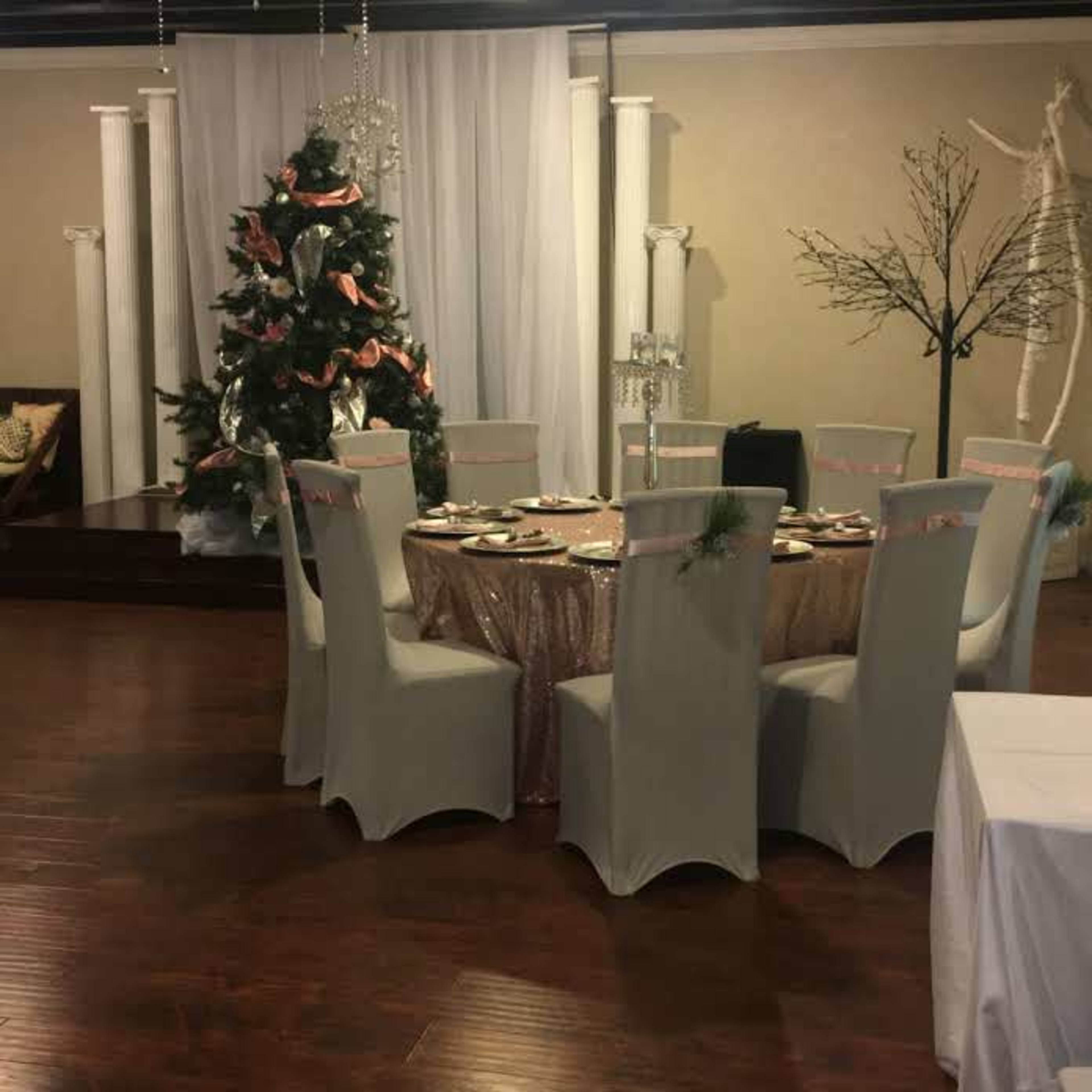 A dining area is set up with a round table draped in a light cloth, surrounded by eight chairs, with a Christmas tree and decorative elements in the background.