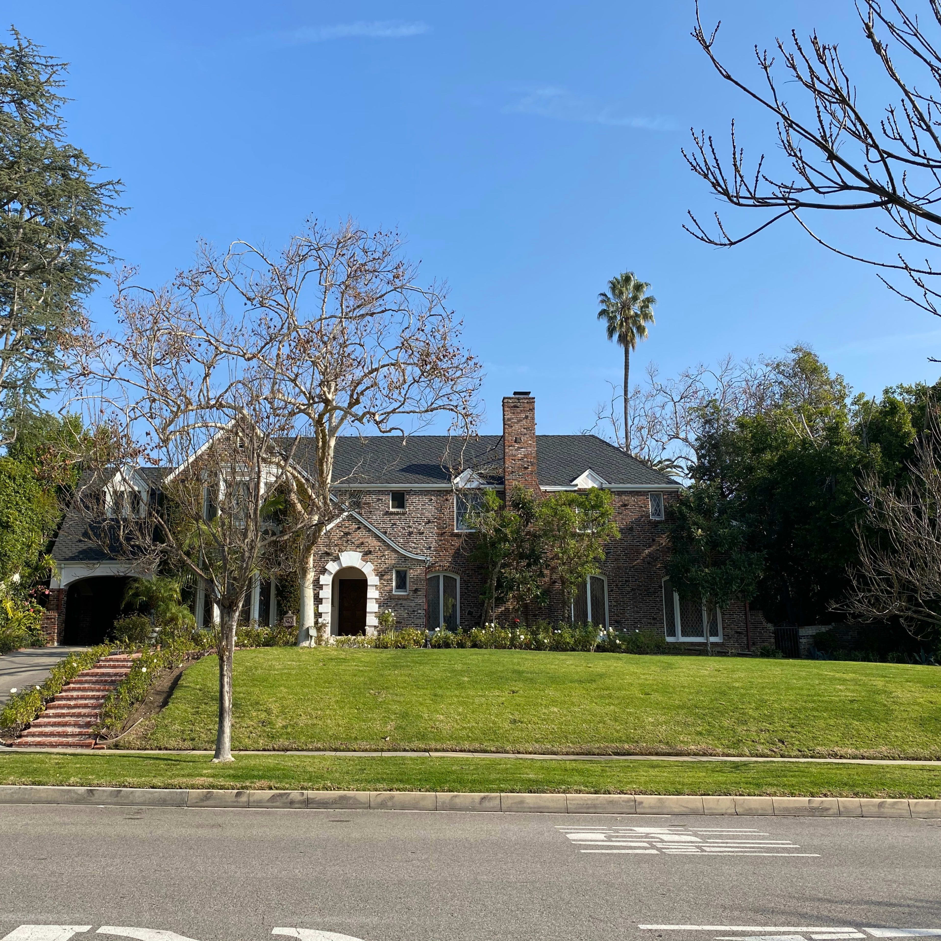 A large brick house with a steep roof is set on a grassy lot lined with trees and a driveway.