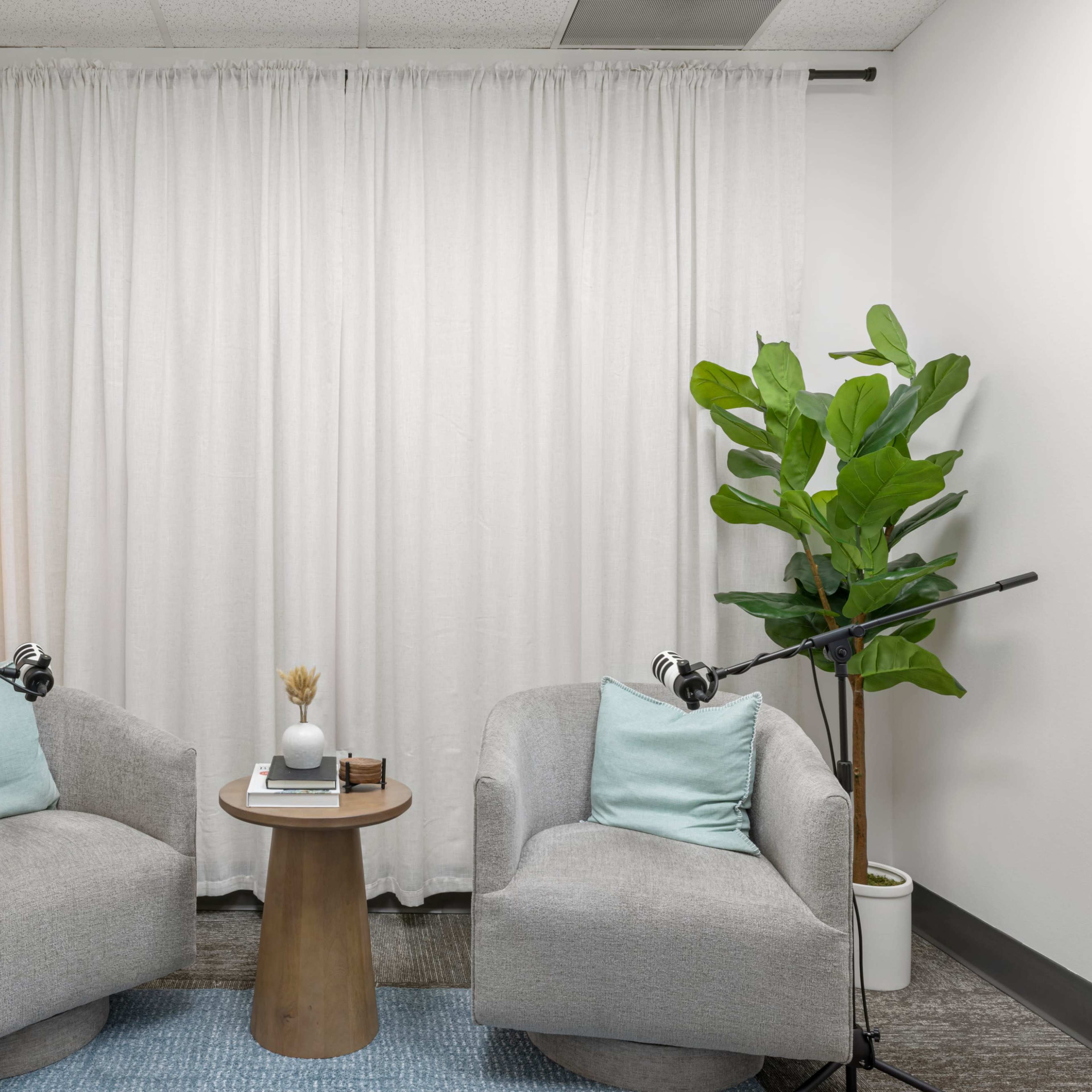 A small, modern space with two gray armchairs, a round wooden table, and a potted plant, all arranged in front of a white curtain.