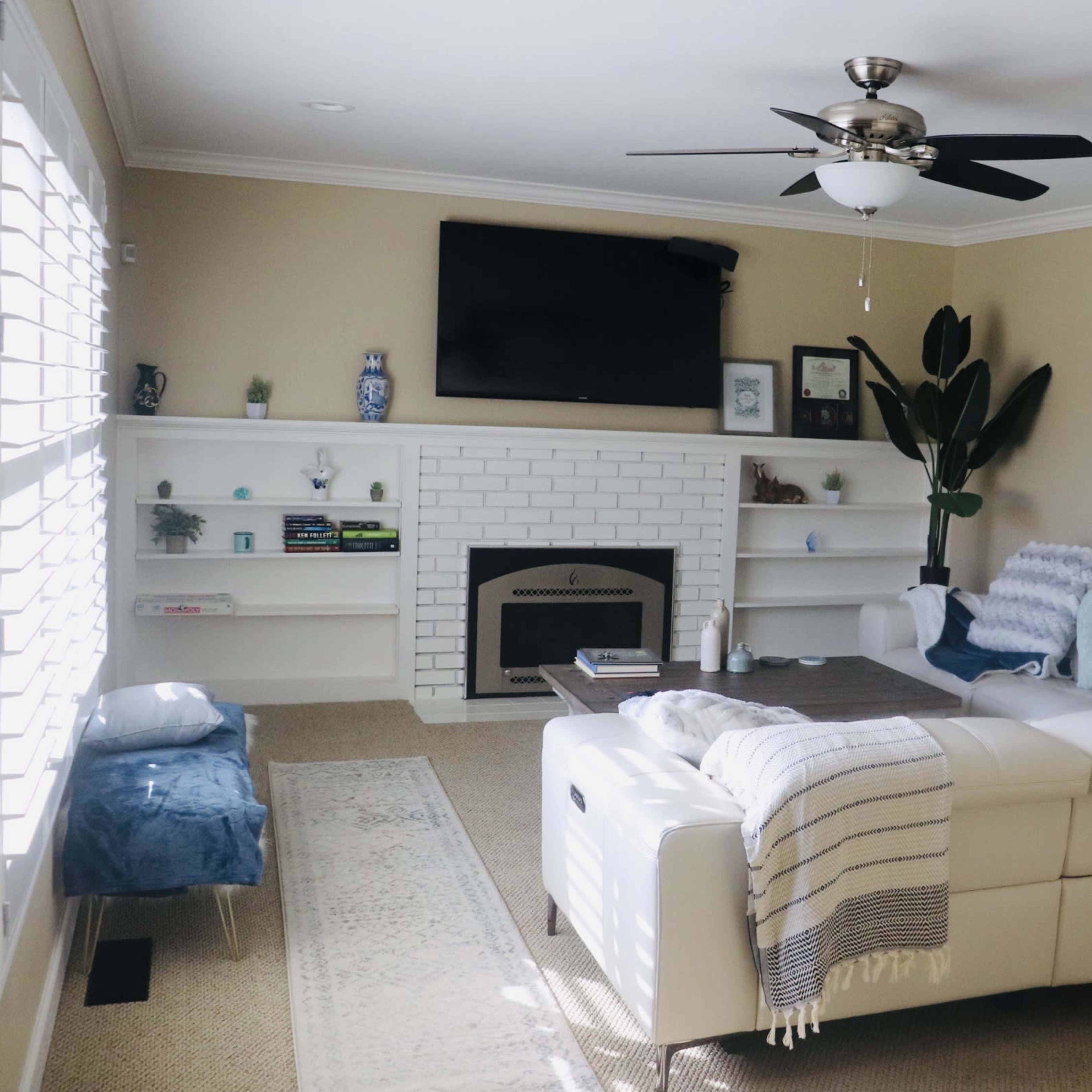 A bright living room features a white sectional sofa, a ceiling fan, a flat-screen TV mounted above a white brick fireplace, and a blue and white artwork on the wall.