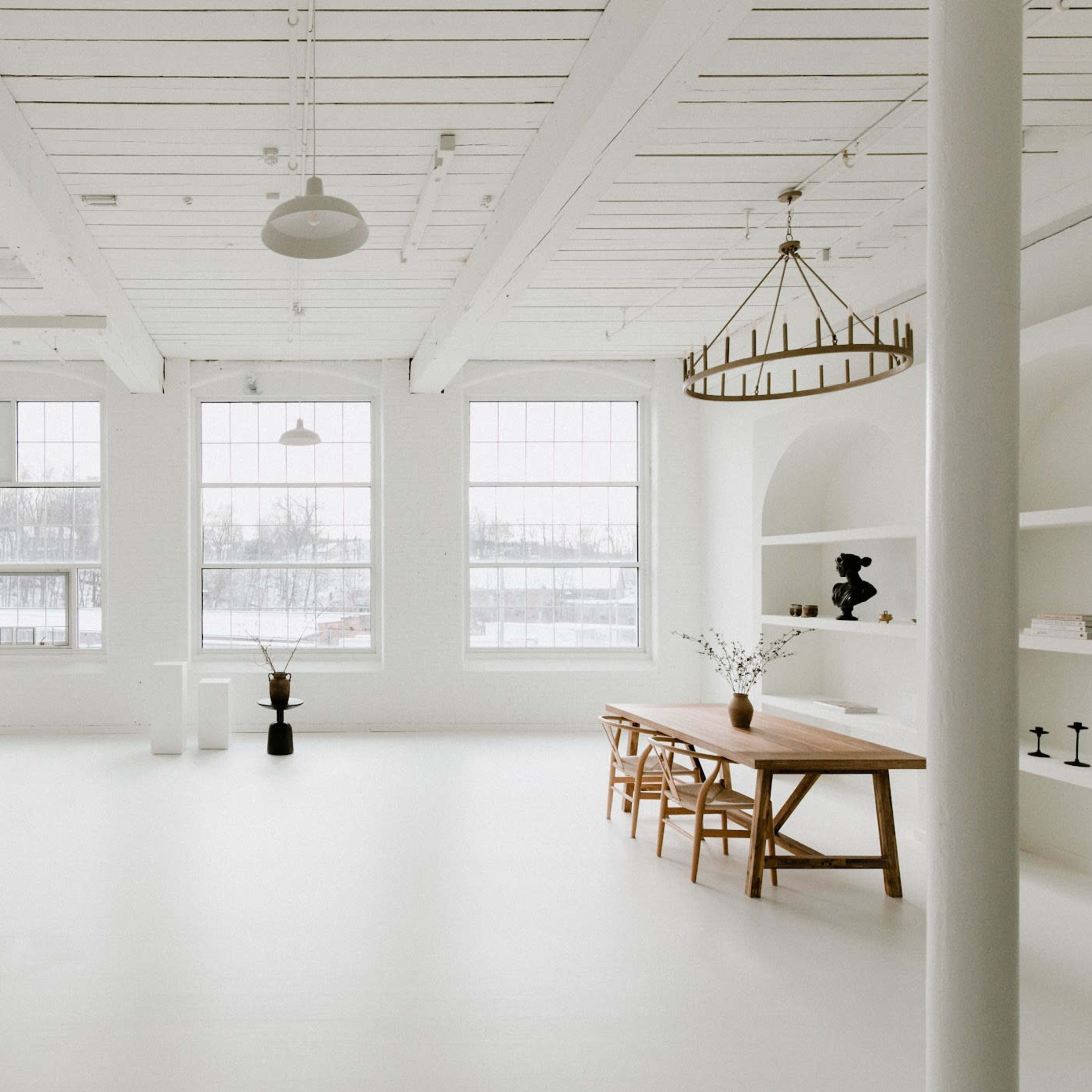 A bright, minimalist interior space featuring a sofa, a wooden dining table, and large windows surrounded by white walls and ceiling beams.