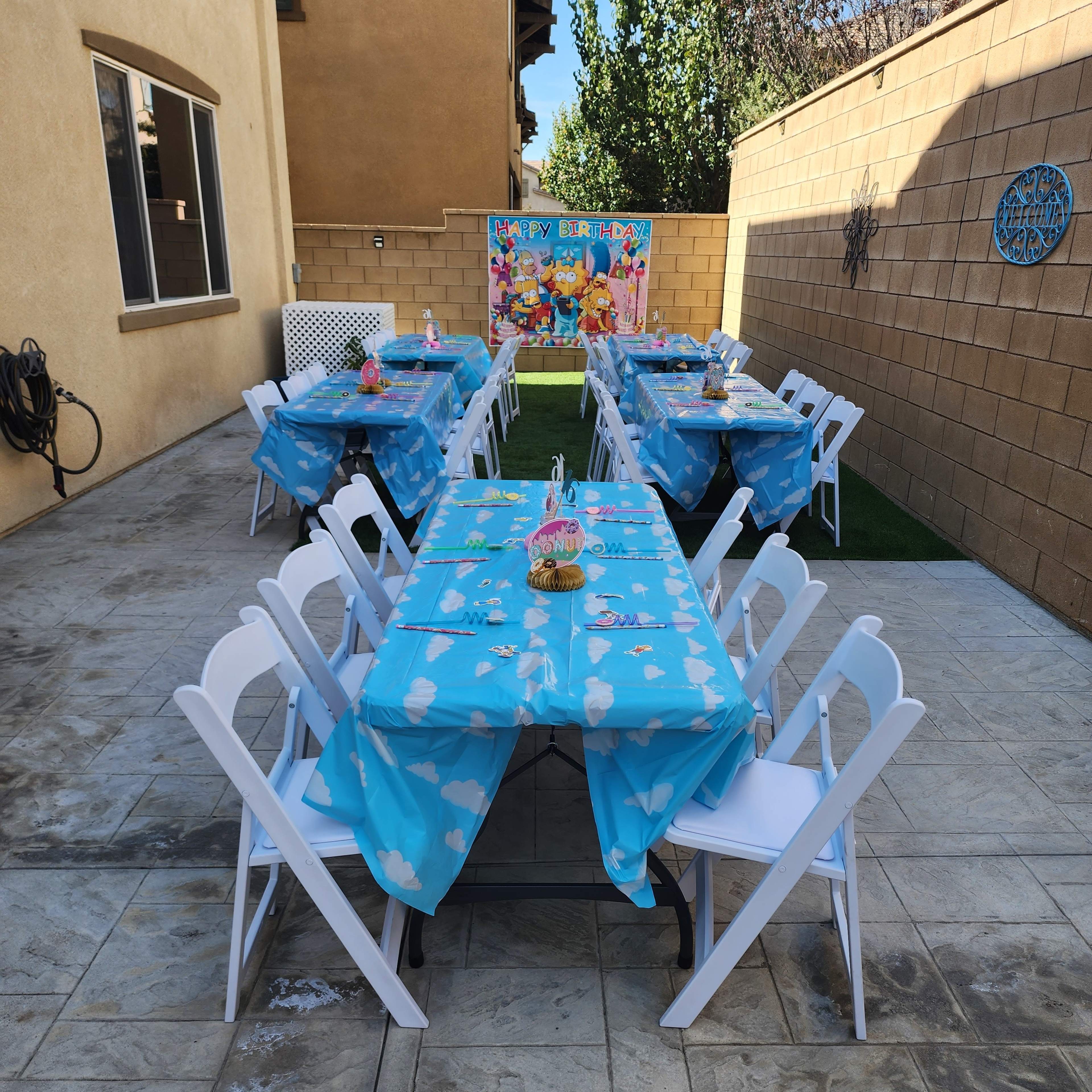 A backyard setup features two rows of tables covered with blue tablecloths, arranged for a birthday celebration, with a colorful backdrop and decorations in the background.