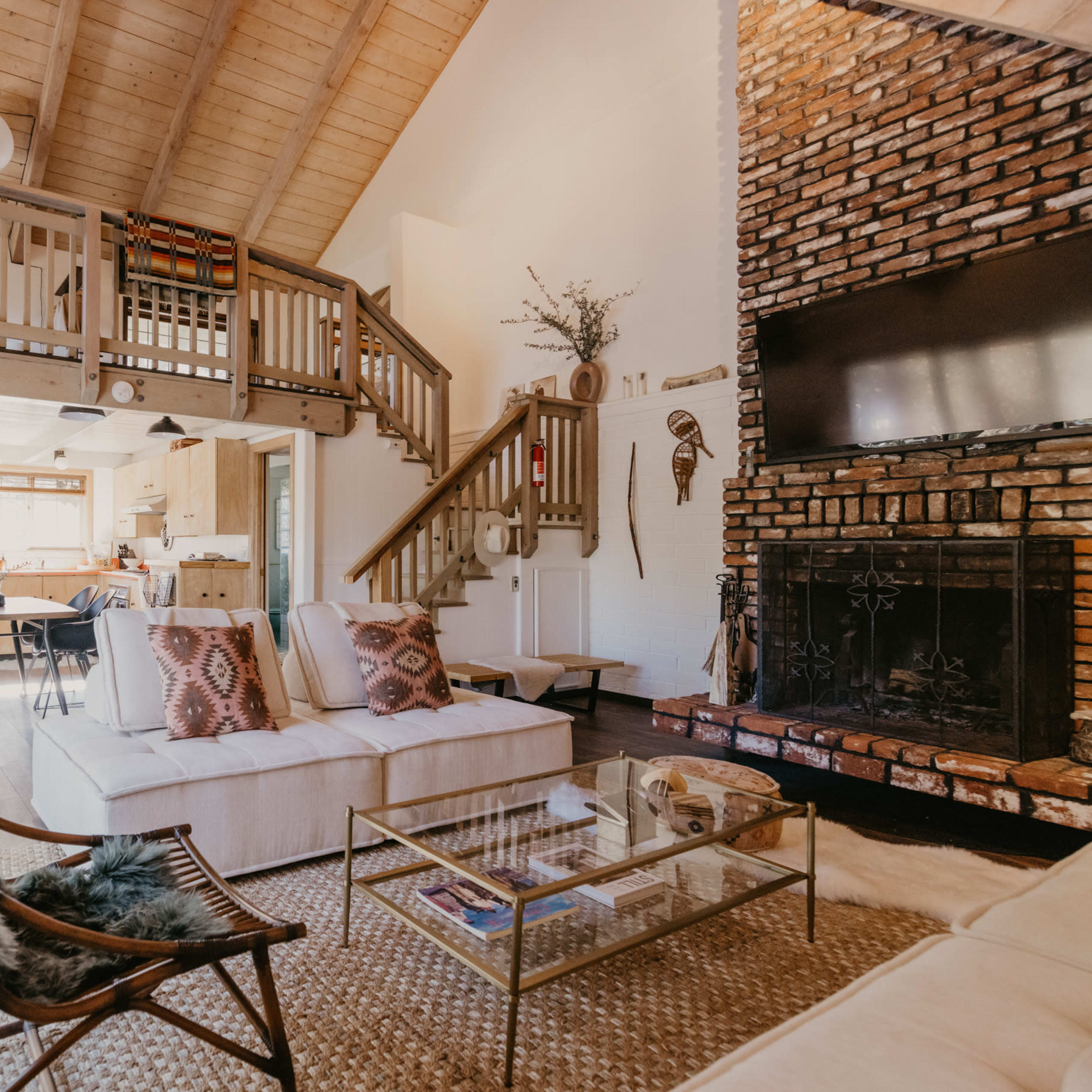 A cozy living room features a white sectional sofa, a glass coffee table, and a large brick fireplace, with a staircase leading to a lofted area above.