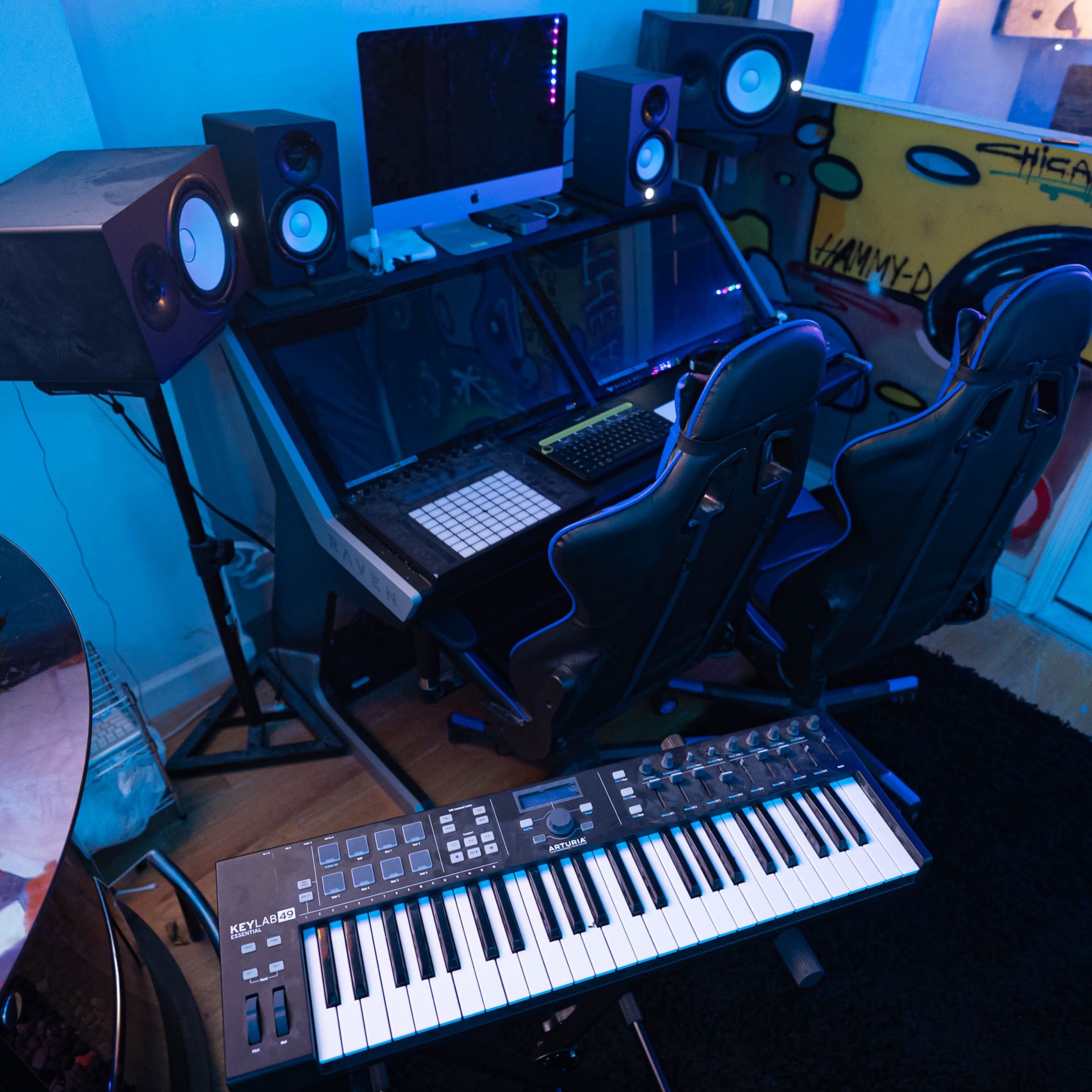 A music production setup featuring a desk with an Apple iMac, two loudspeakers, a keyboard controller on the floor, and a dark rug under the equipment.