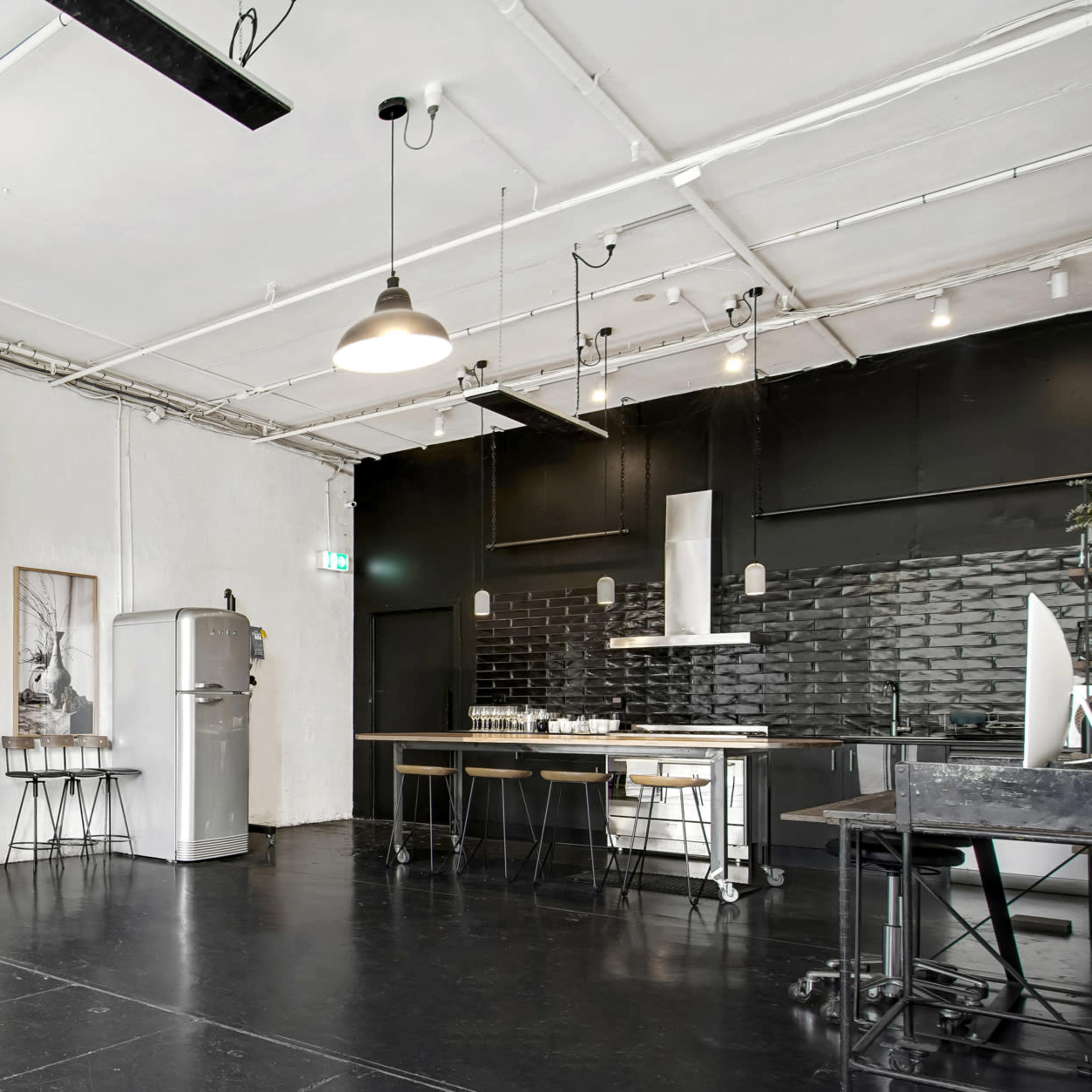 A spacious, minimalist room featuring a blank white wall, a dark kitchen area with a refrigerator, and a large black tabletop.
