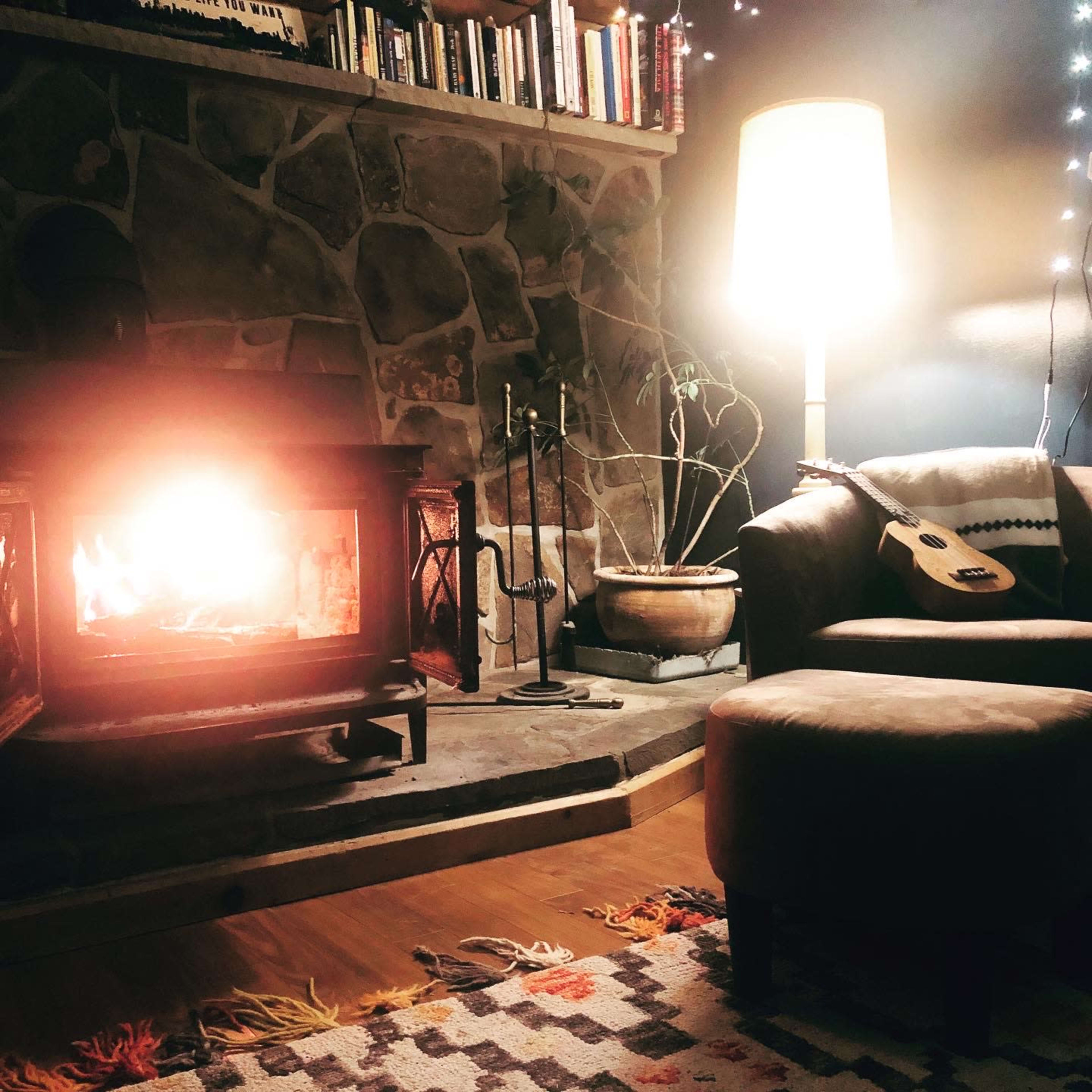 A cozy living room features a stone fireplace with a lit fire, a comfortable chair and ottoman, and a lamp illuminating the space.