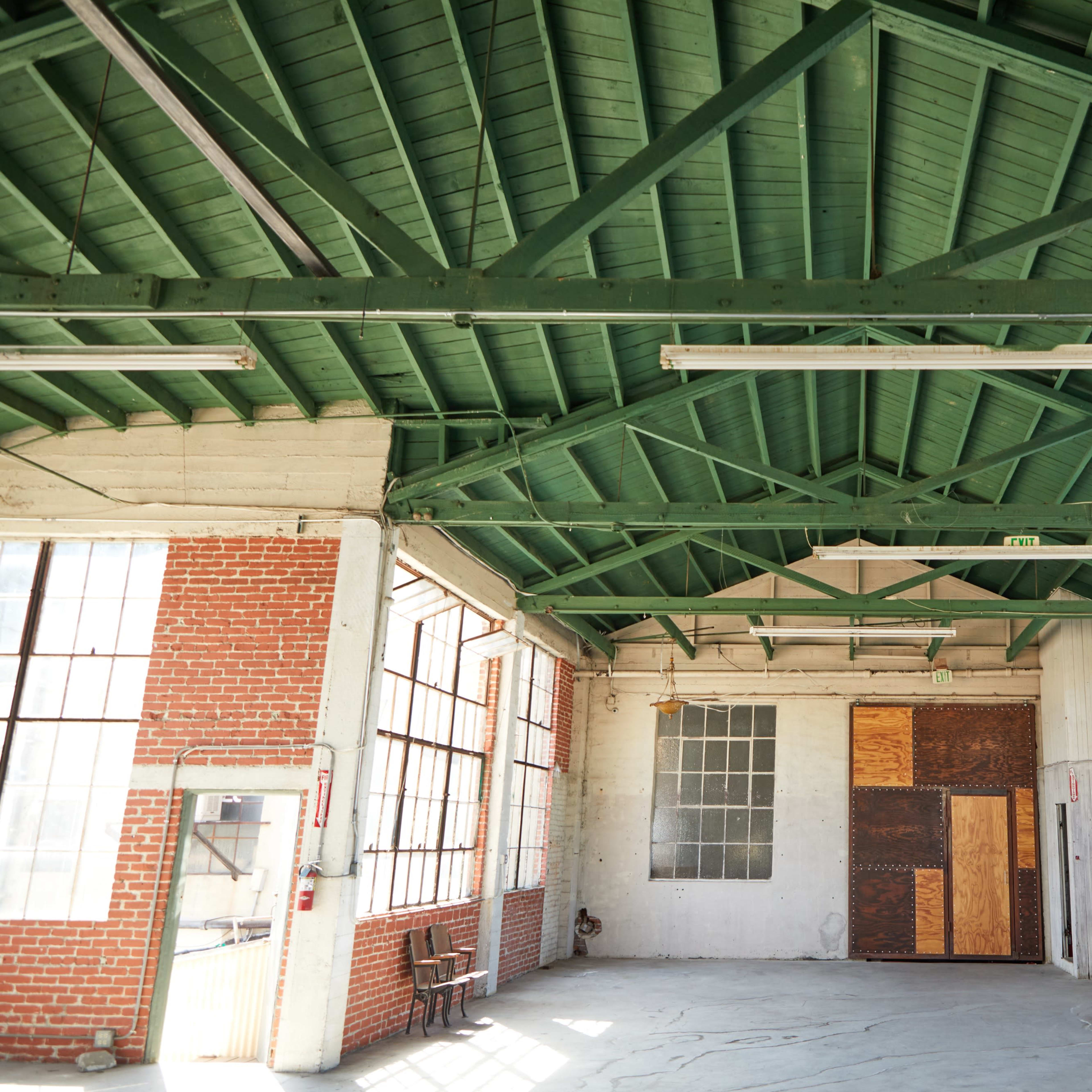 An empty industrial space with a green wooden ceiling, brick walls, and large windows allowing natural light to enter.