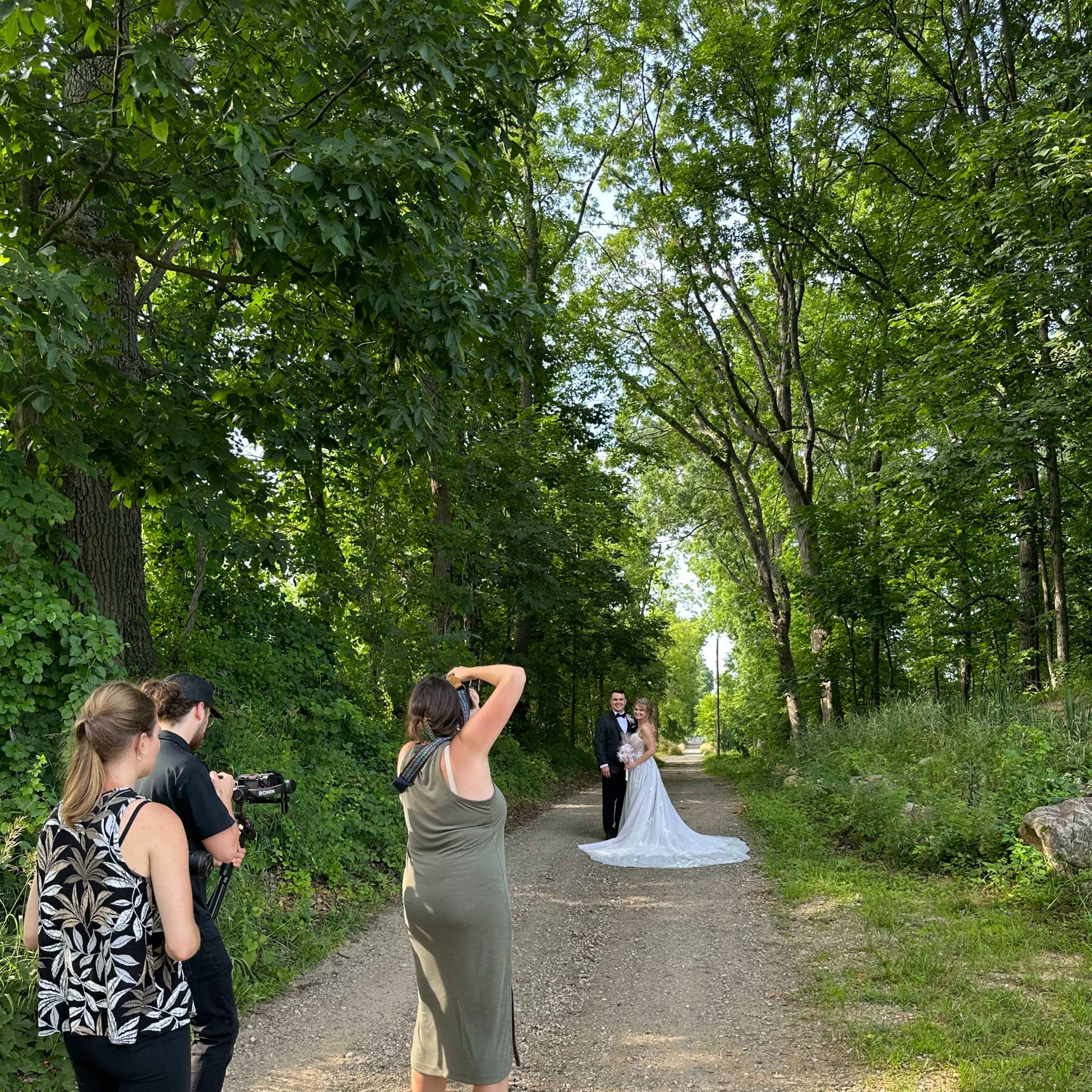 A bride and groom stand on a dirt path surrounded by trees while a photographer captures their portrait.