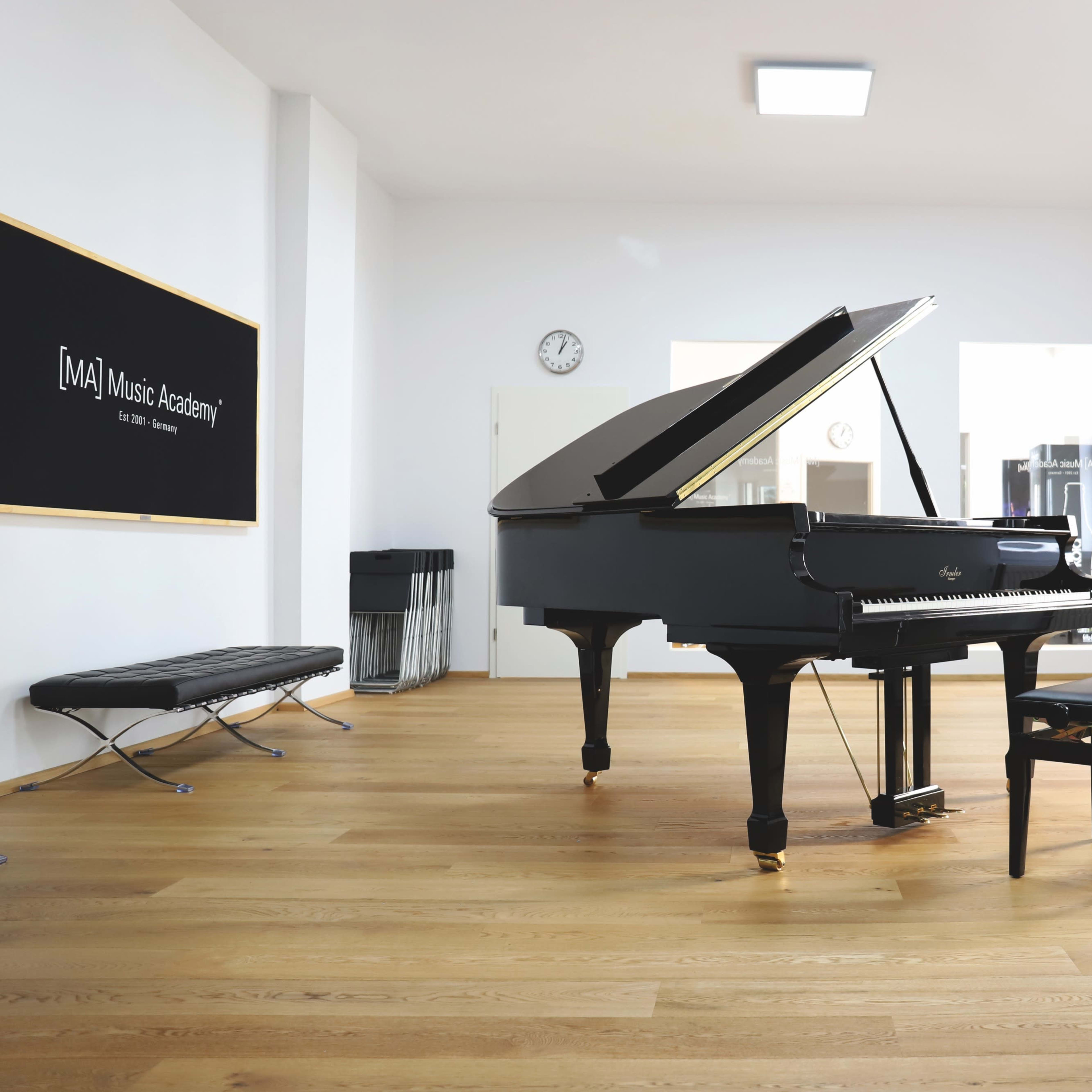 A grand piano is set in a spacious, well-lit music studio with wooden flooring and a black bench beside it.