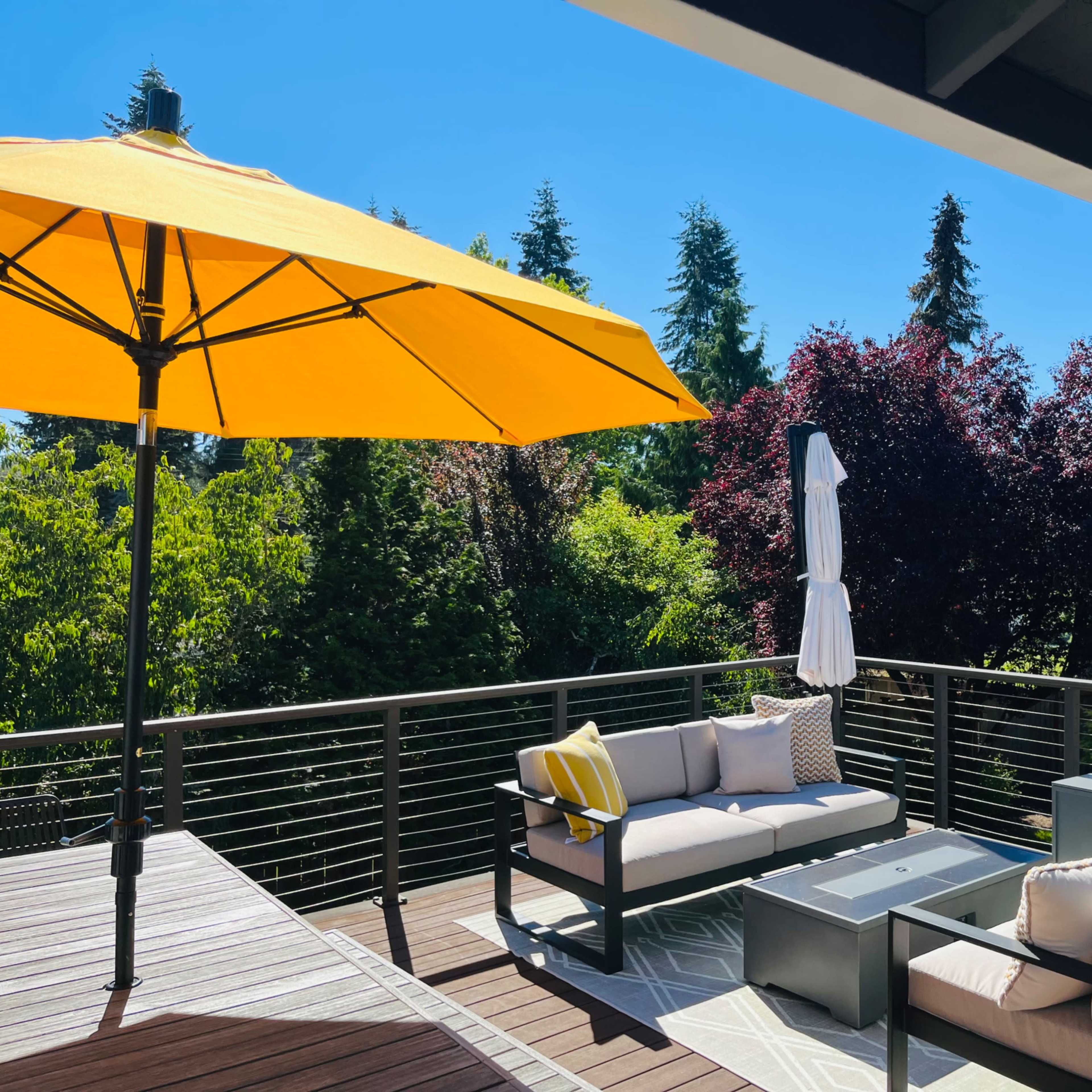 A deck featuring a yellow umbrella, grey seating with cushions, and surrounding lush trees.