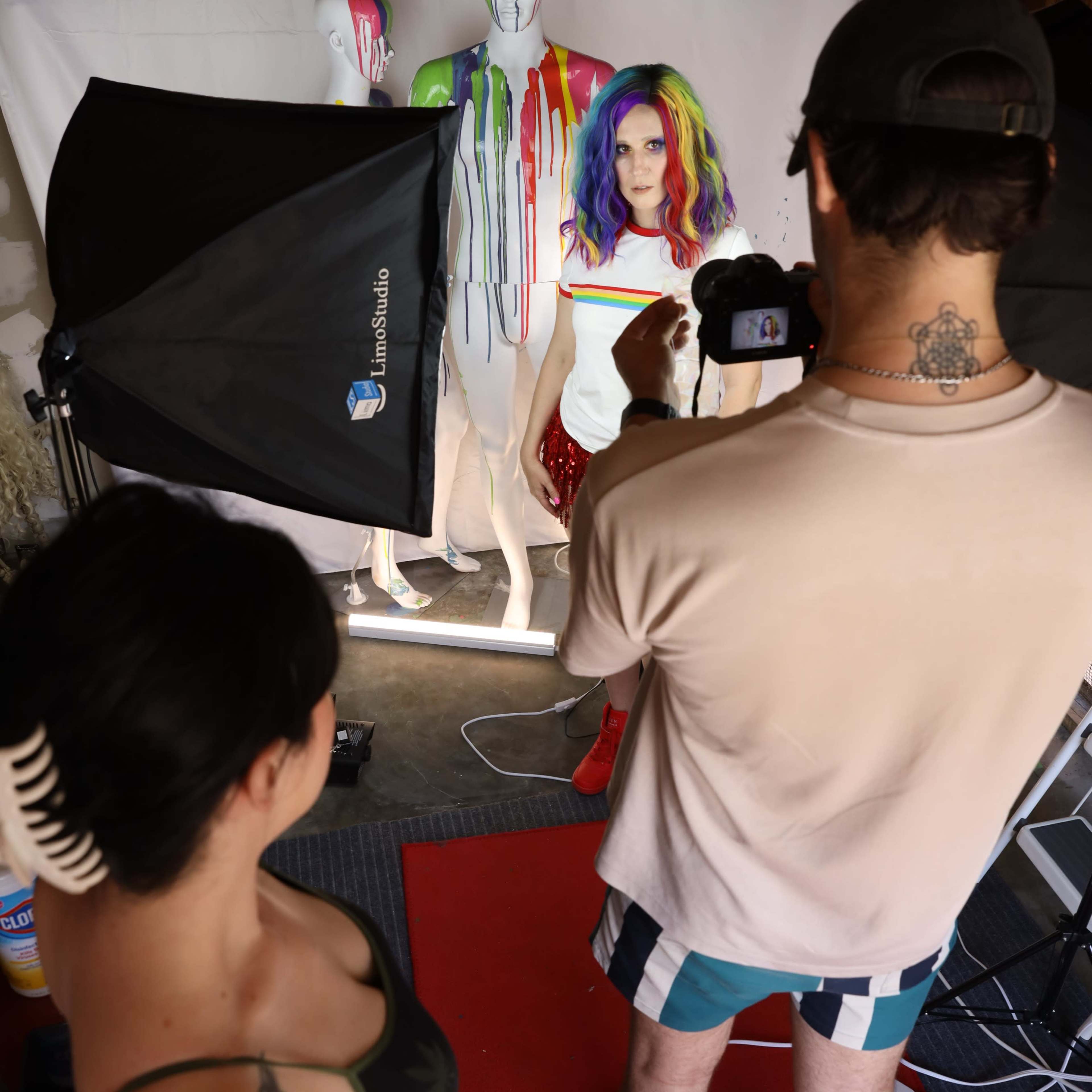 A model with colorful hair poses in front of painted mannequins while a photographer captures the scene in a studio setting.