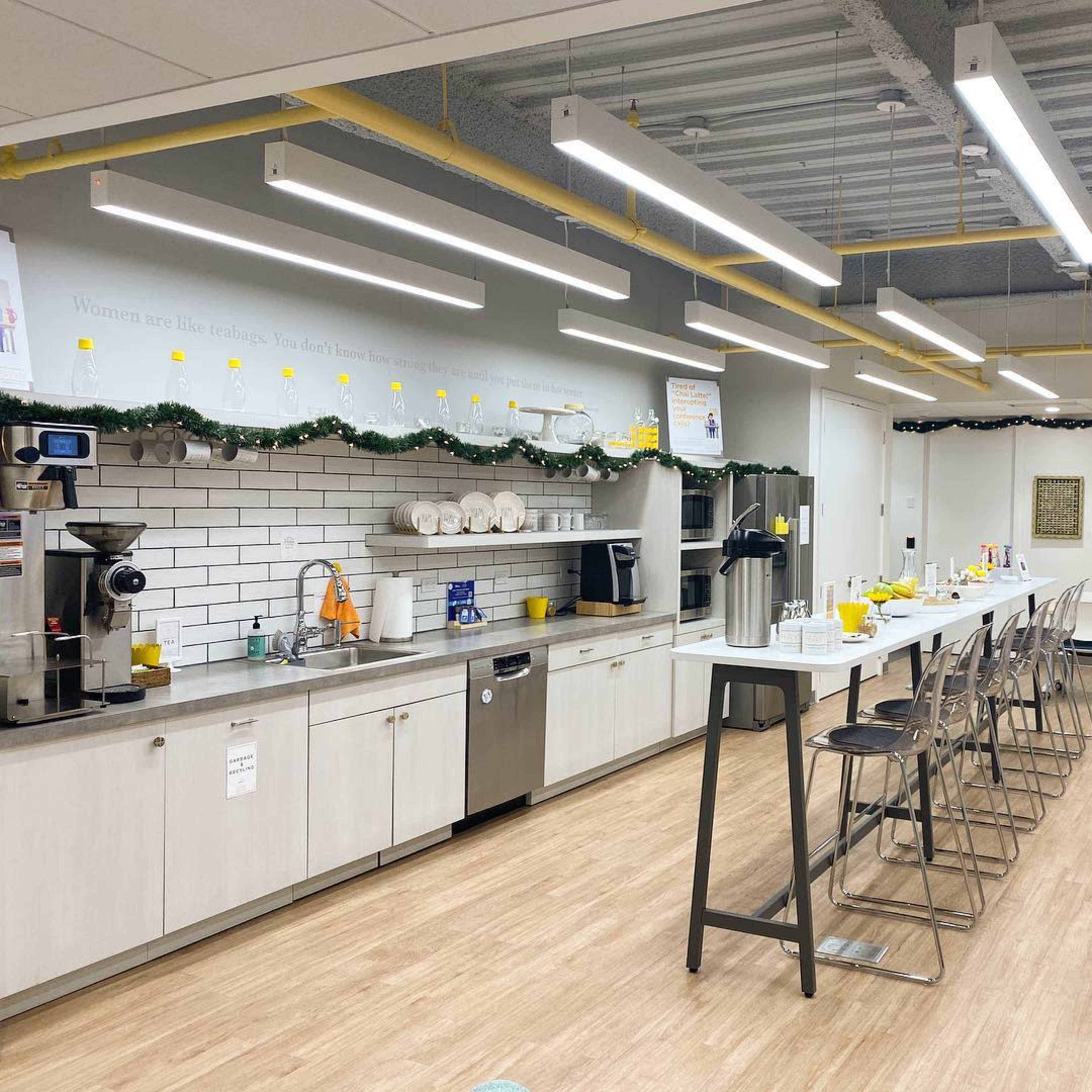 A modern kitchen and dining area features a long countertop with bar stools, a coffee machine, and neatly arranged dishes, all under bright overhead lights.