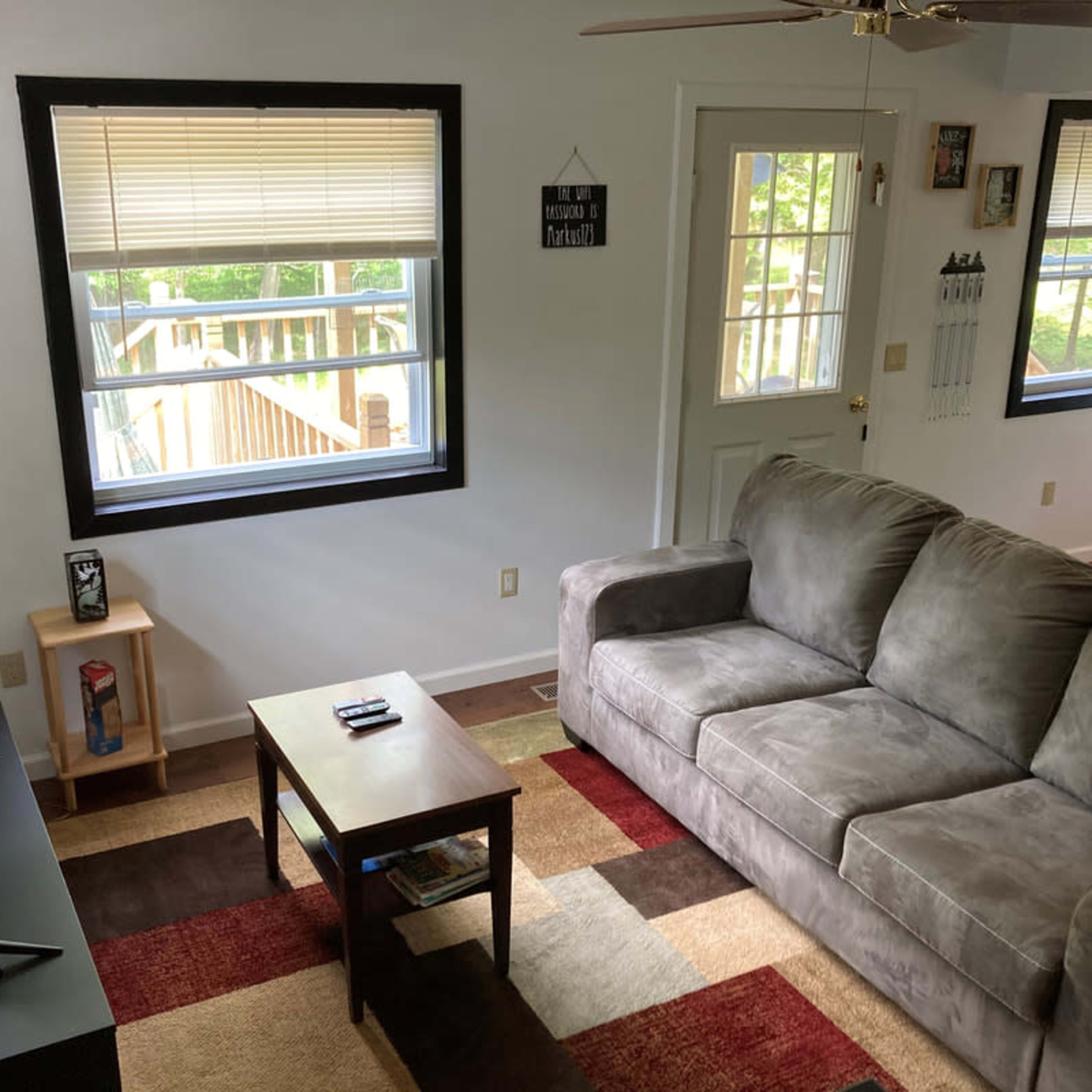 A living room with a beige couch, a coffee table, a television on a stand, and two windows facing a wooden deck outside.