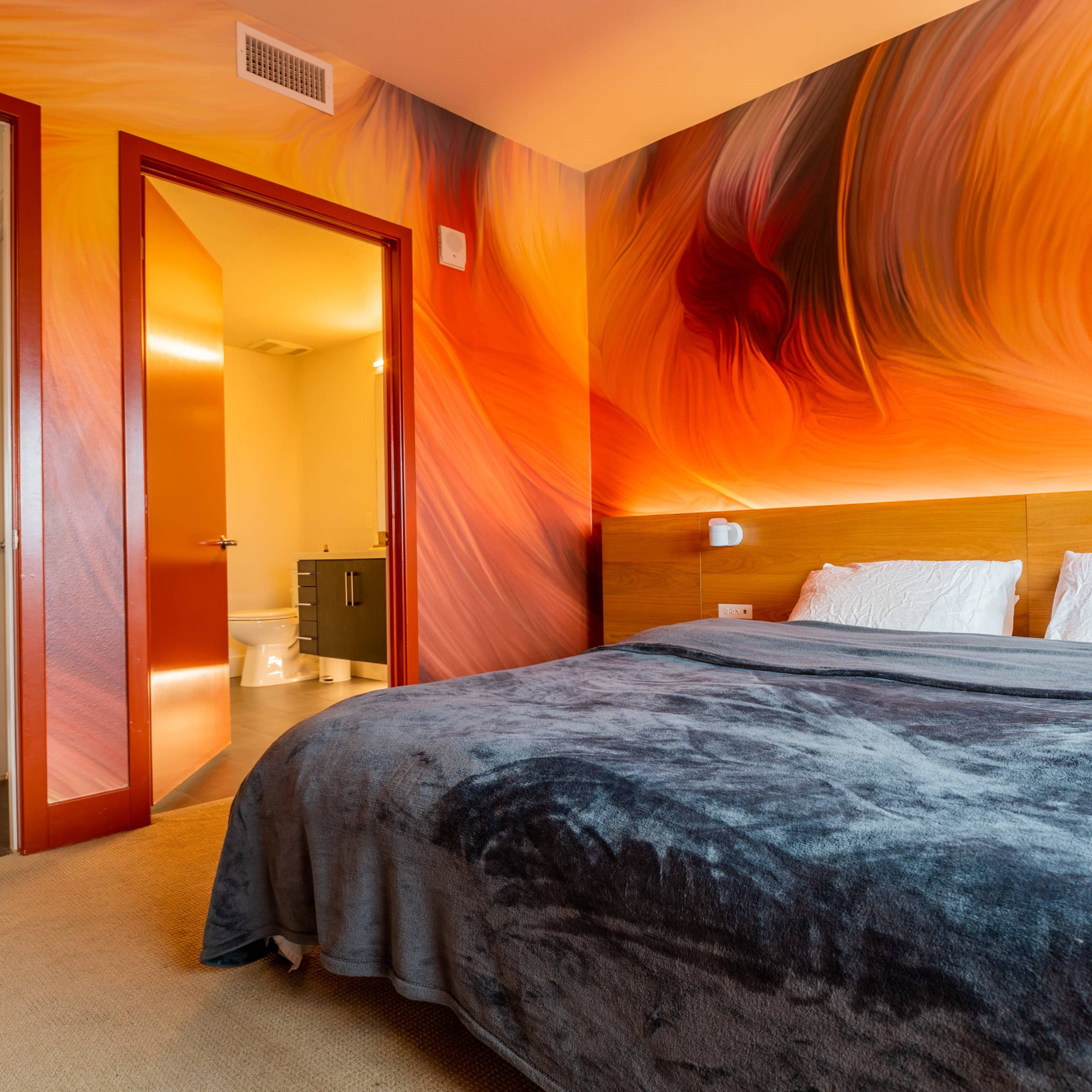 A bedroom with a large bed covered in a gray blanket, featuring a vibrant orange and red swirl mural on the wall, and a doorway leading to a bathroom in the background.