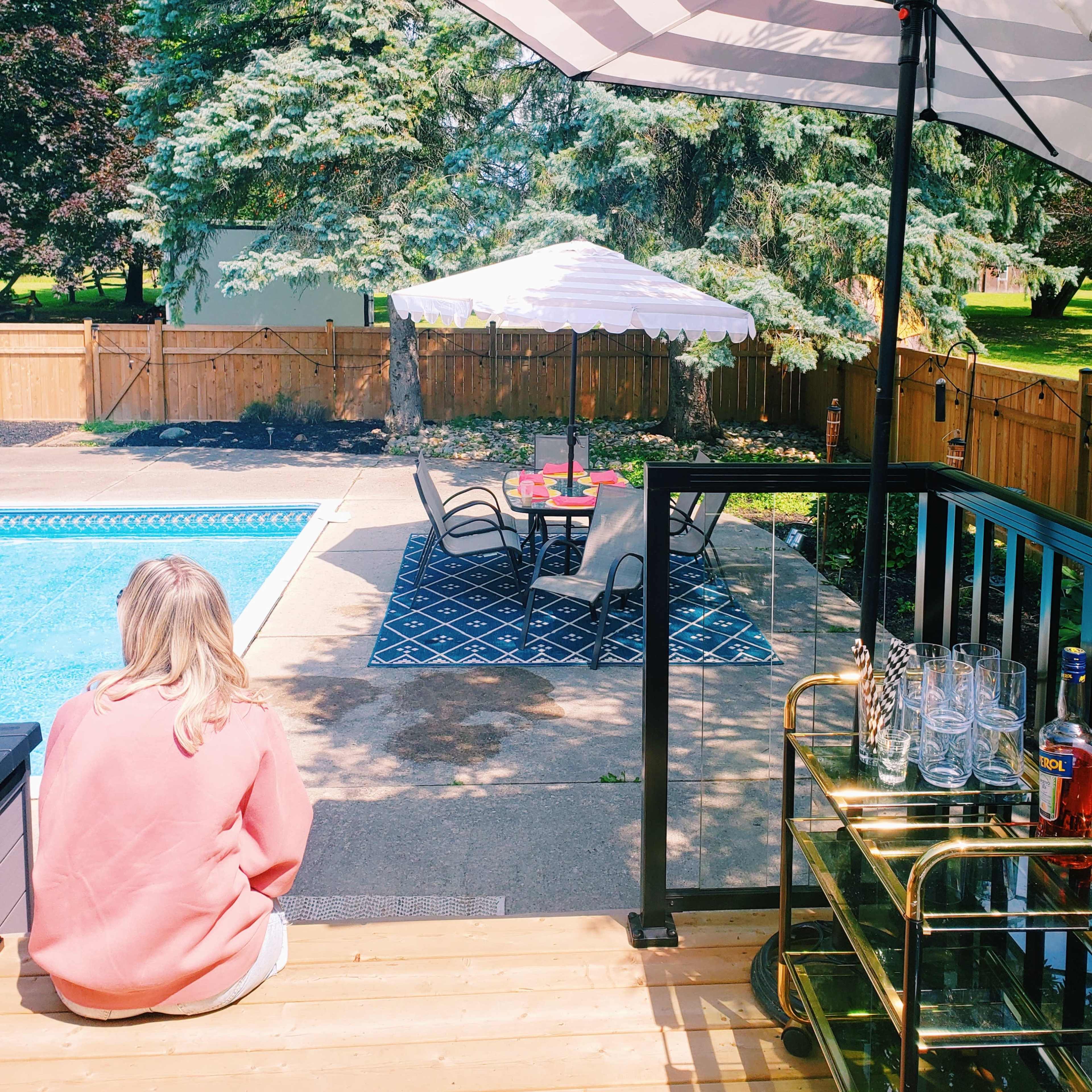 A person sits on a wooden deck near a swimming pool, overlooking a table set with an umbrella and chairs in a backyard.