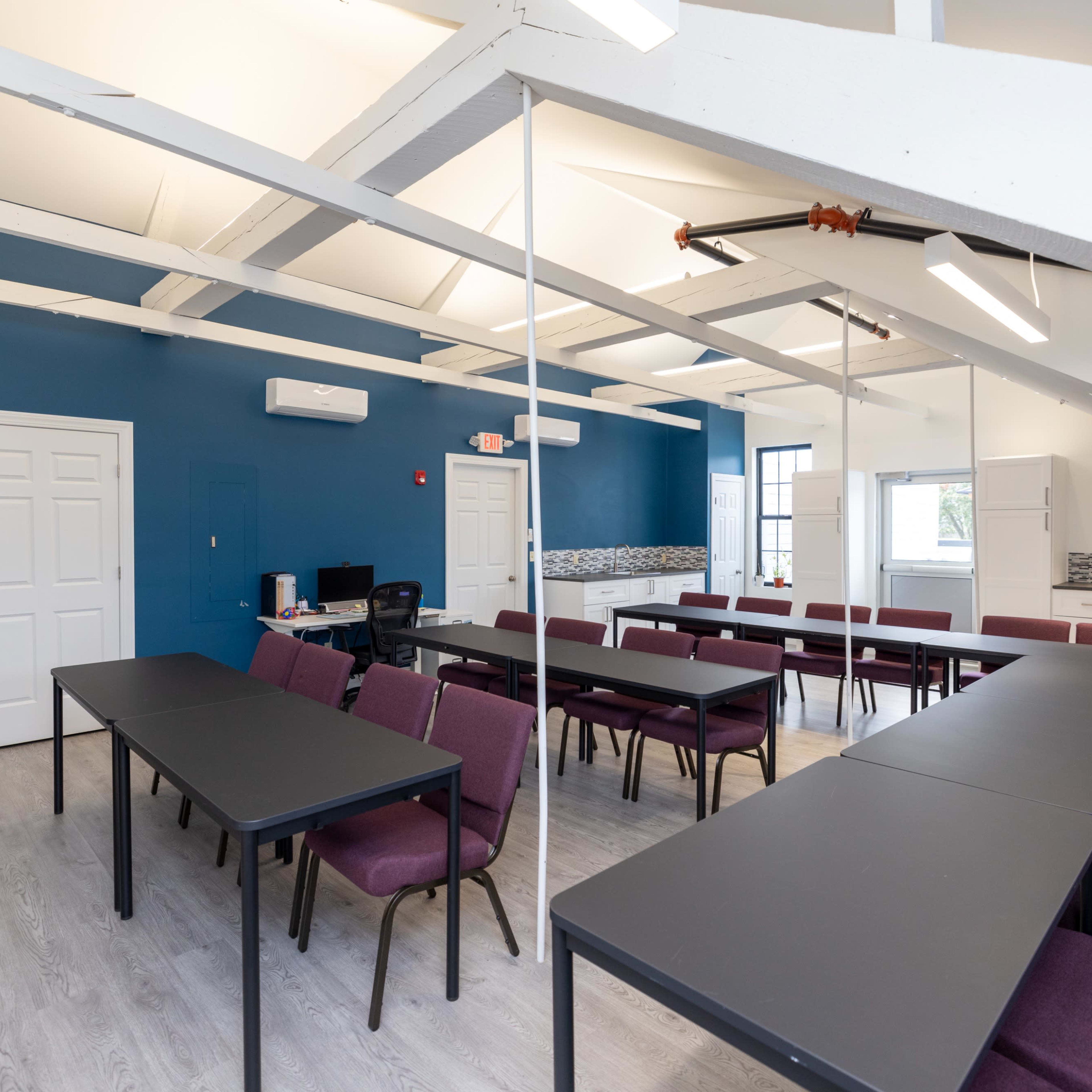 A bright, spacious conference room with several tables arranged in a U-shape and chairs positioned around them, featuring blue walls and large windows.
