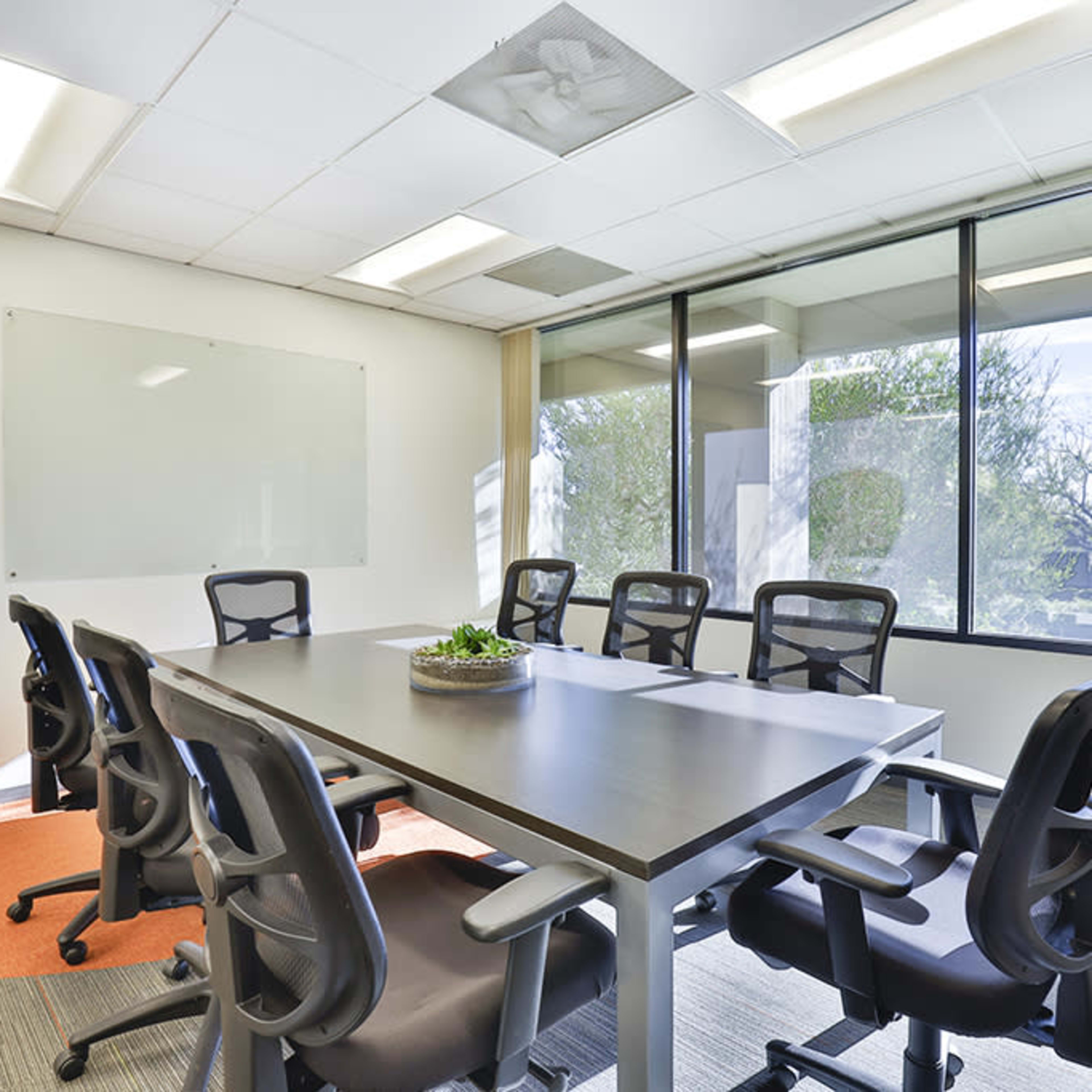 A modern conference room features a large table surrounded by ergonomic chairs, with a glass wall offering views of the outdoors and a plant in the corner.