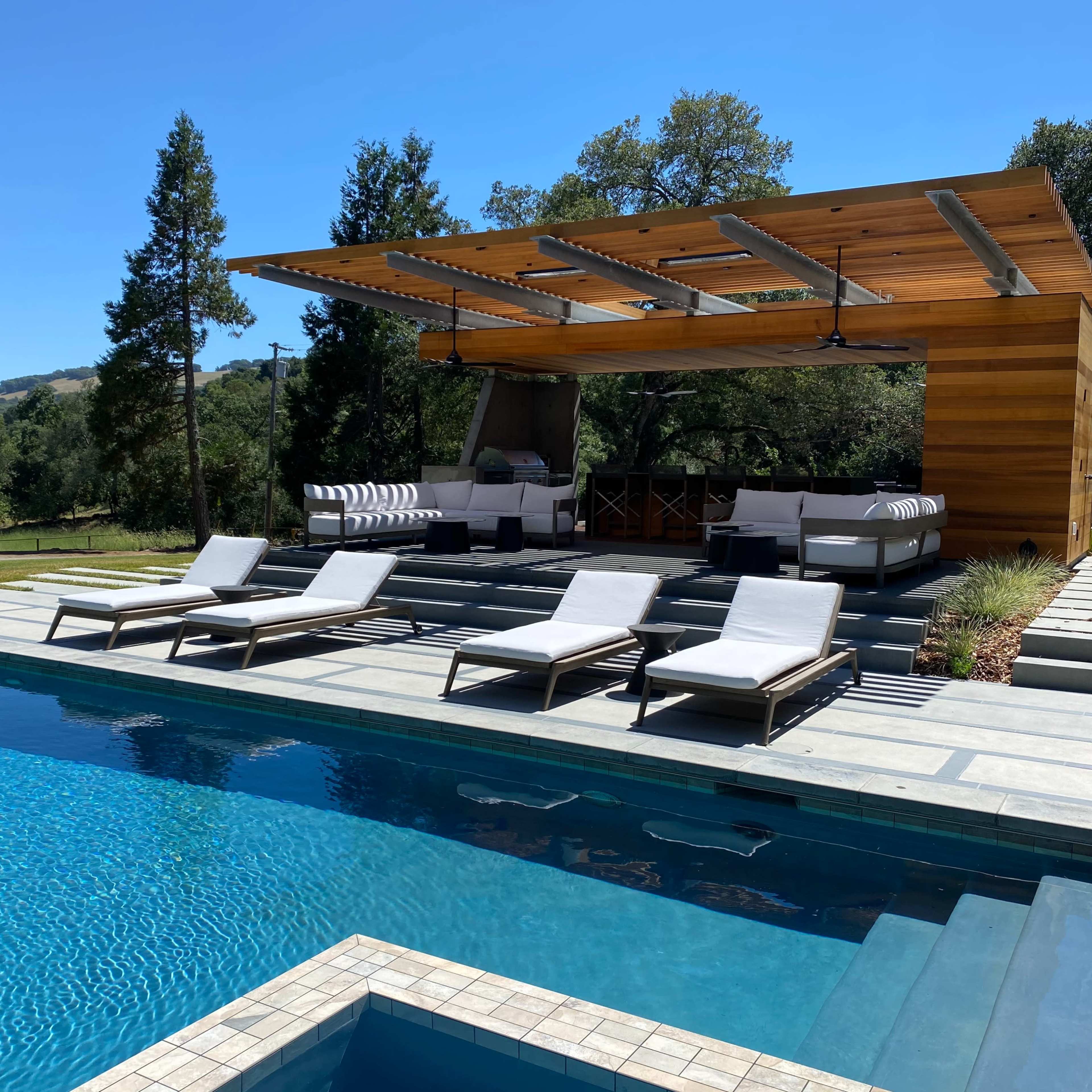A modern outdoor lounge area with a wooden pergola, poolside loungers, and a swimming pool surrounded by greenery.
