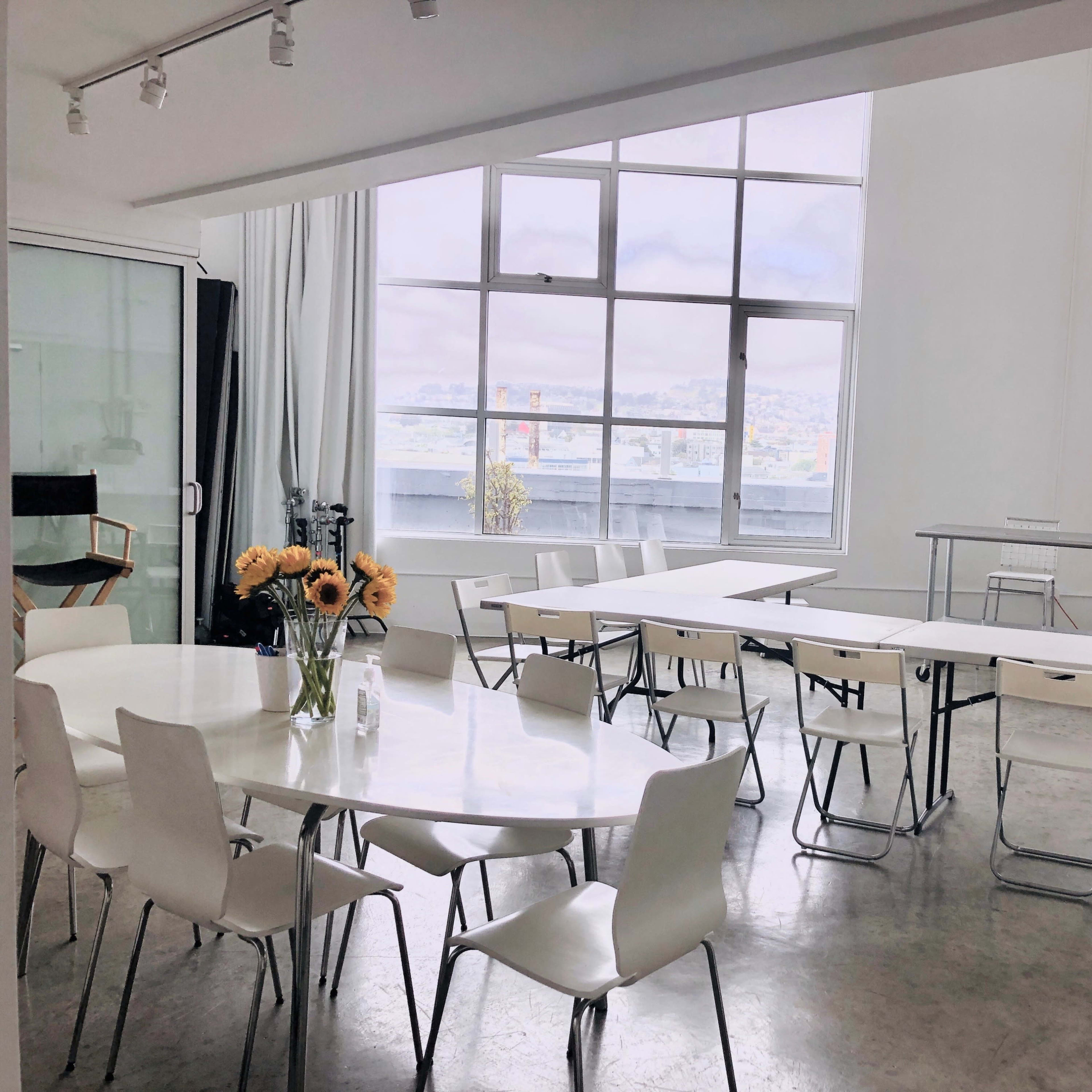 A spacious room features several white tables arranged with chairs and large windows offering a view of the city.