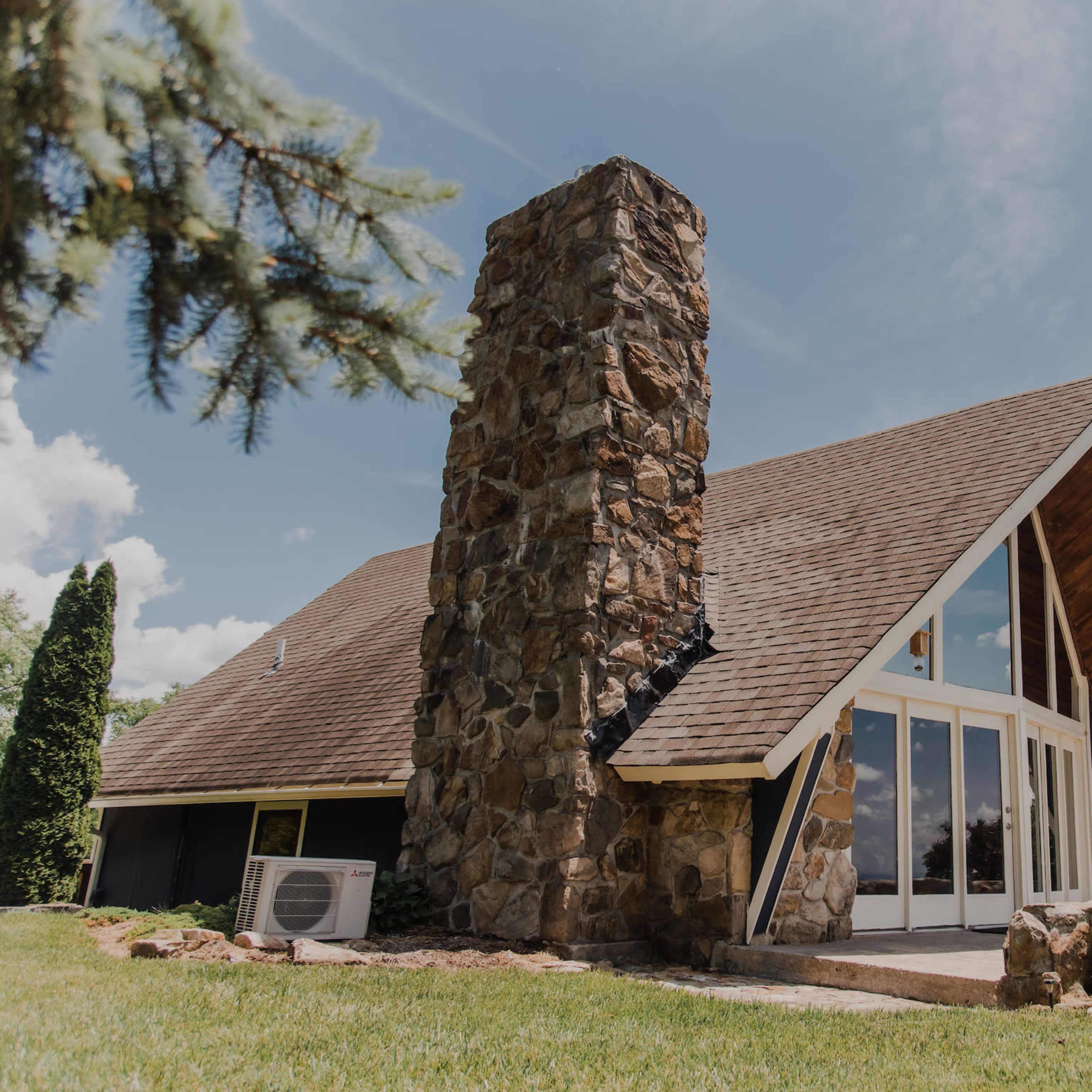 A modern house with a stone chimney, large windows, and a sloped roof, surrounded by green grass and trees.