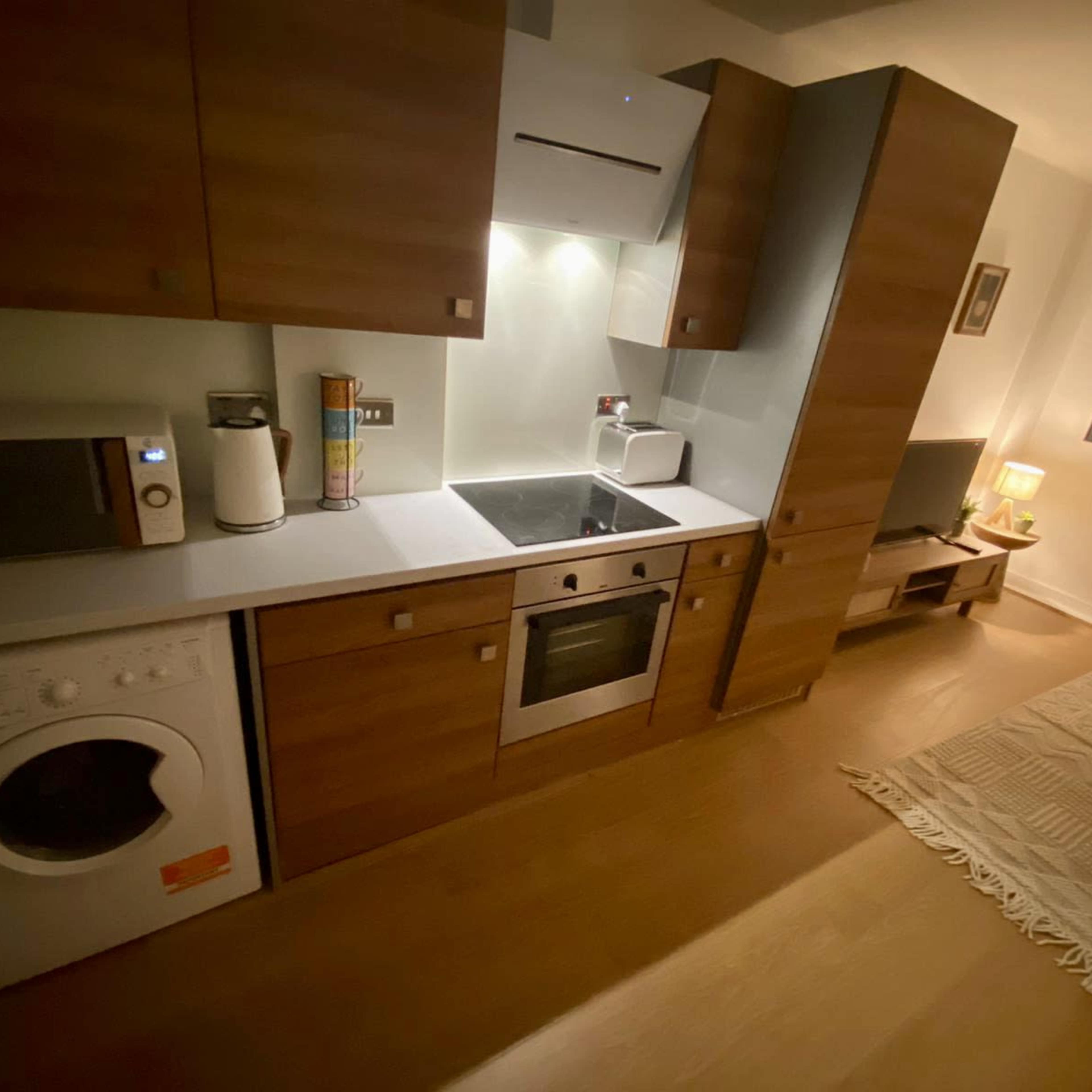 A modern kitchen area with wooden cabinetry, a washing machine, microwave, oven, and countertop integration, adjacent to a living space with a lamp and chair.