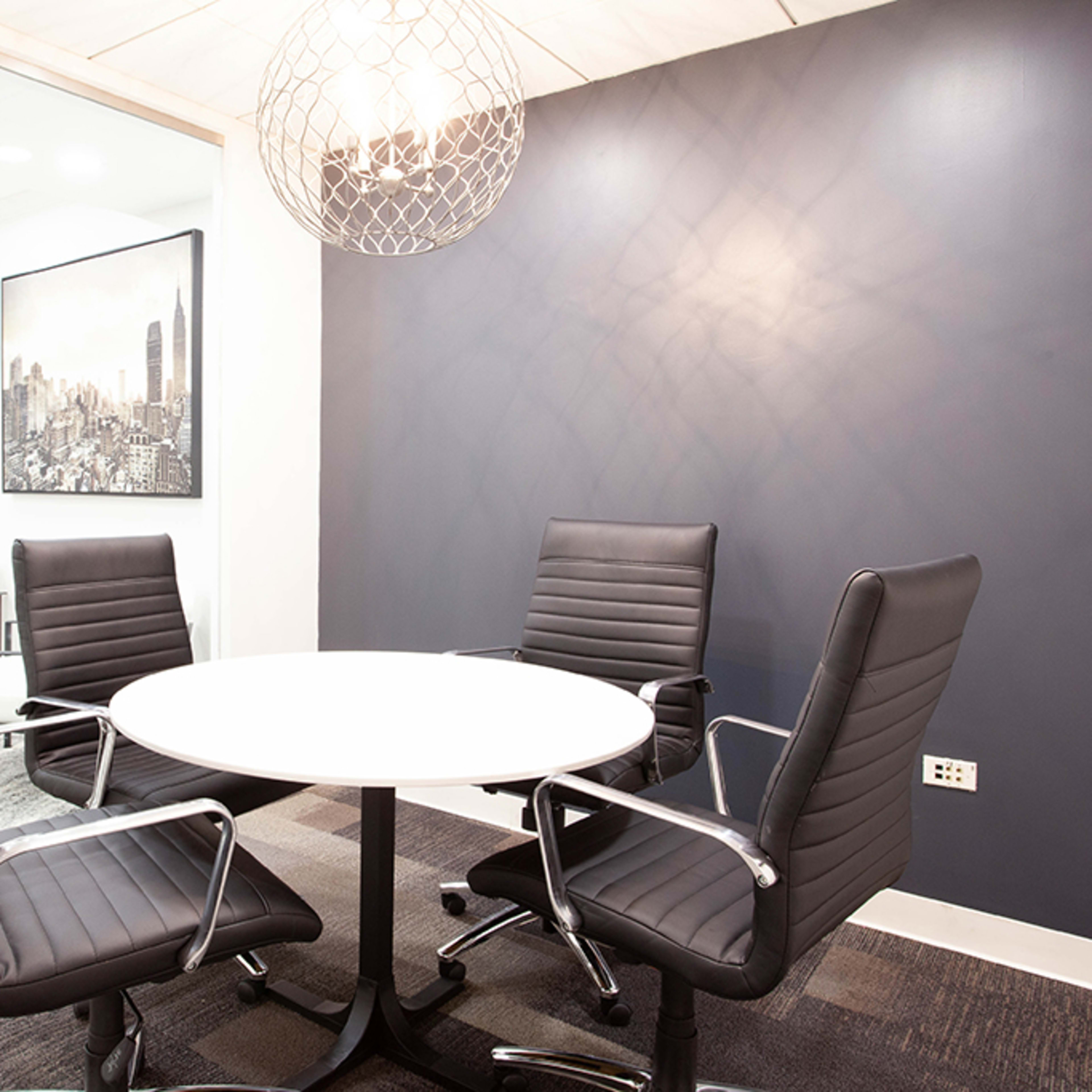 A meeting room features a round white table with four black chairs and a wall-mounted monitor, surrounded by glass walls.