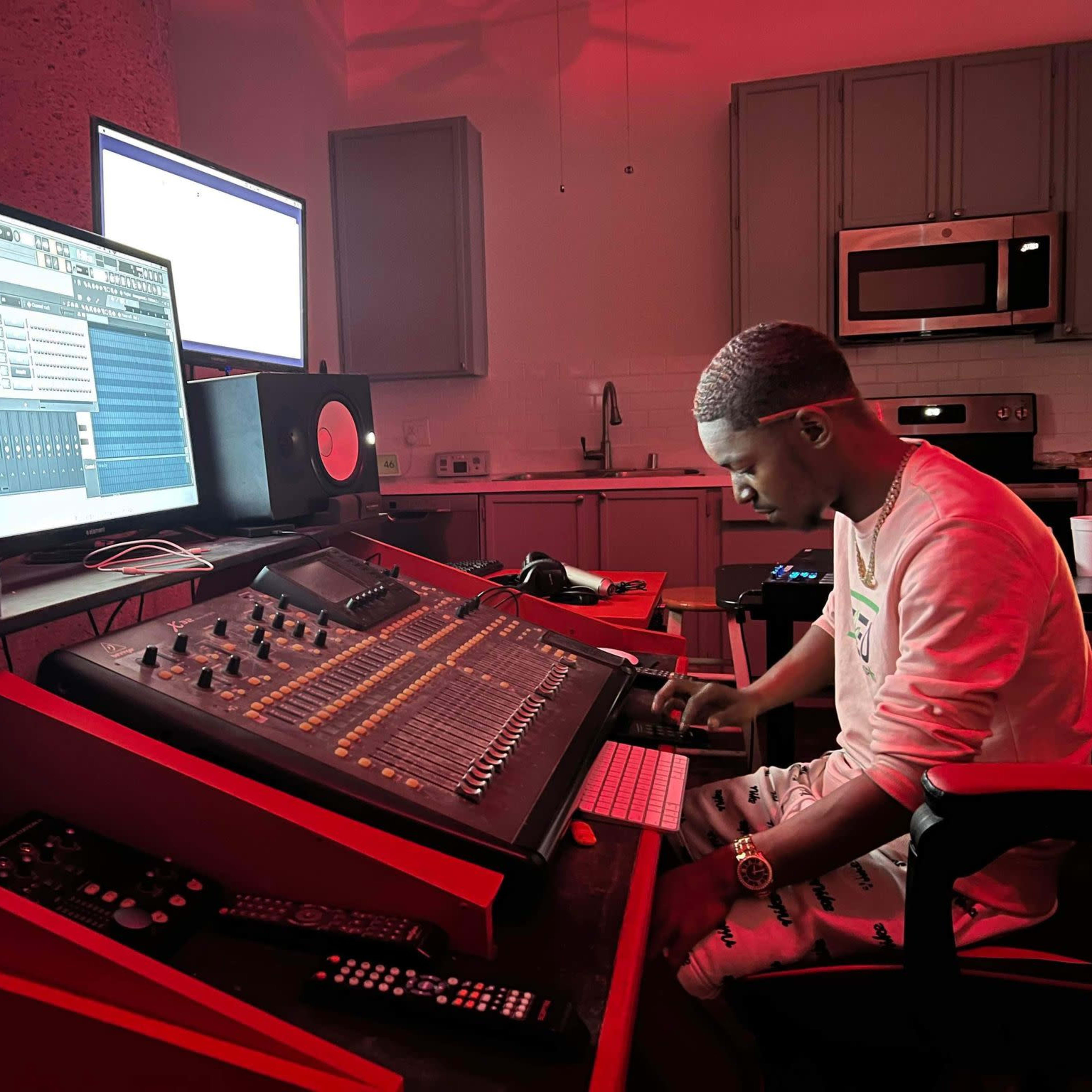 A person is seated at a music production workstation, adjusting controls on a large mixing console in a dimly lit room.