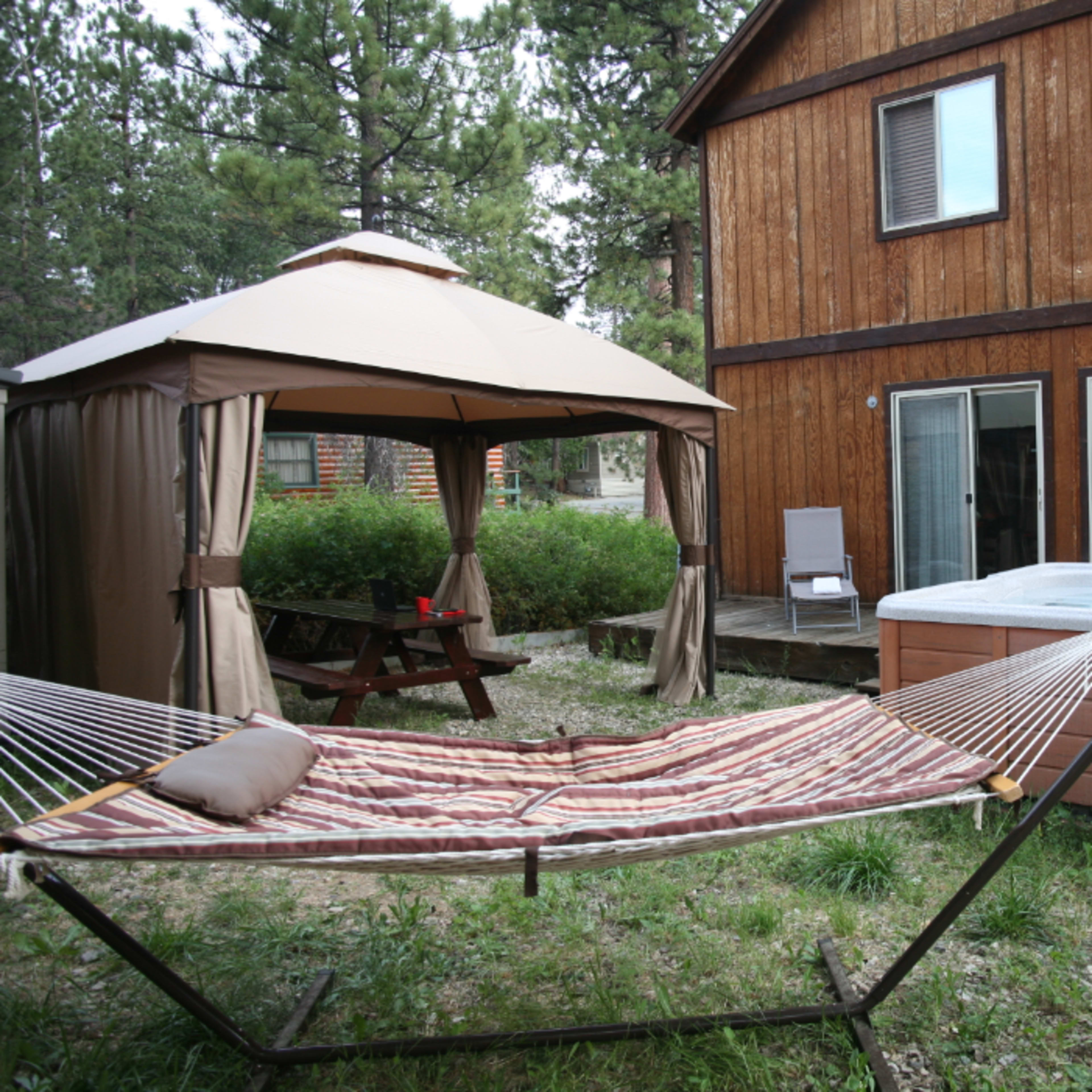 A backyard area features a hammock, a gazebo, a picnic table, and a hot tub next to a wooden house.