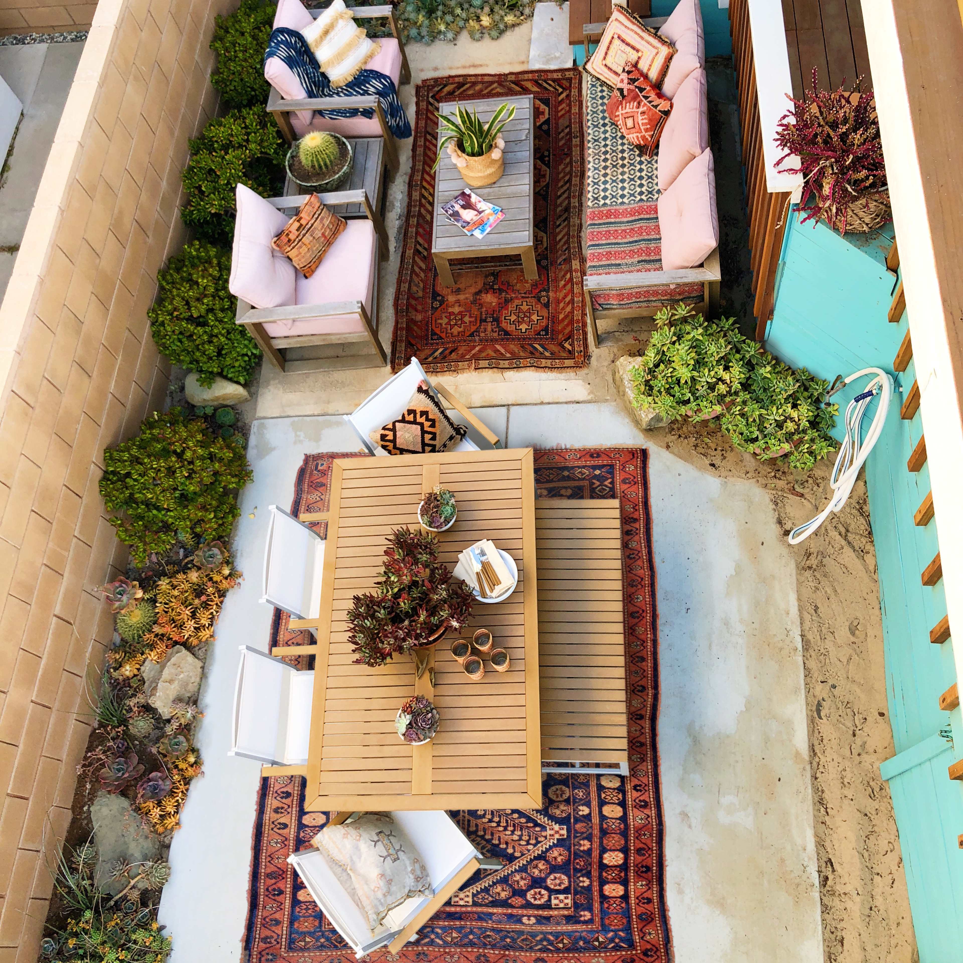 A cozy outdoor seating area featuring a table with chairs, two separate seating arrangements, and various plants, all set on patterned rugs.