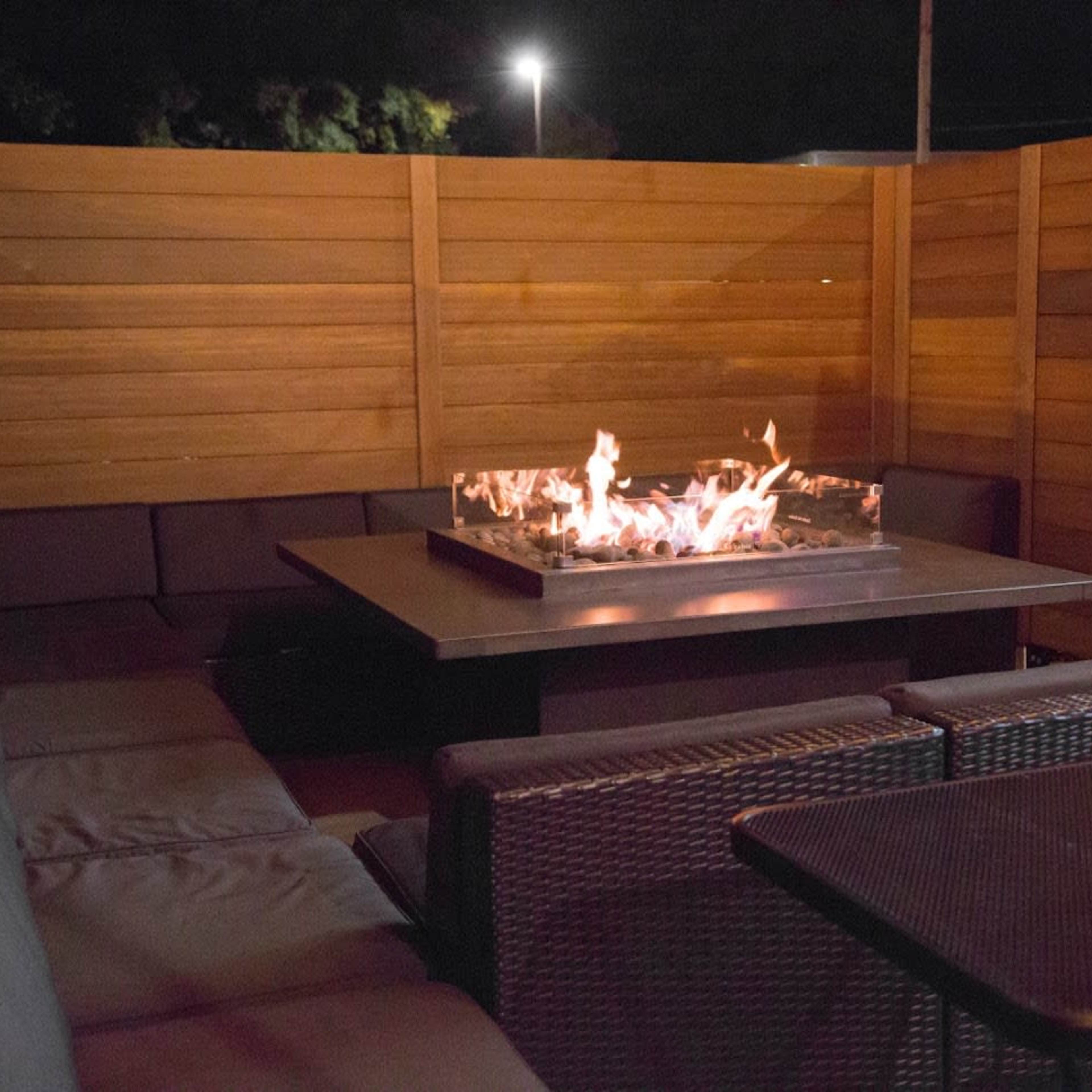 A fire pit surrounded by cushioned seating is situated on a patio enclosed by wooden panels.
