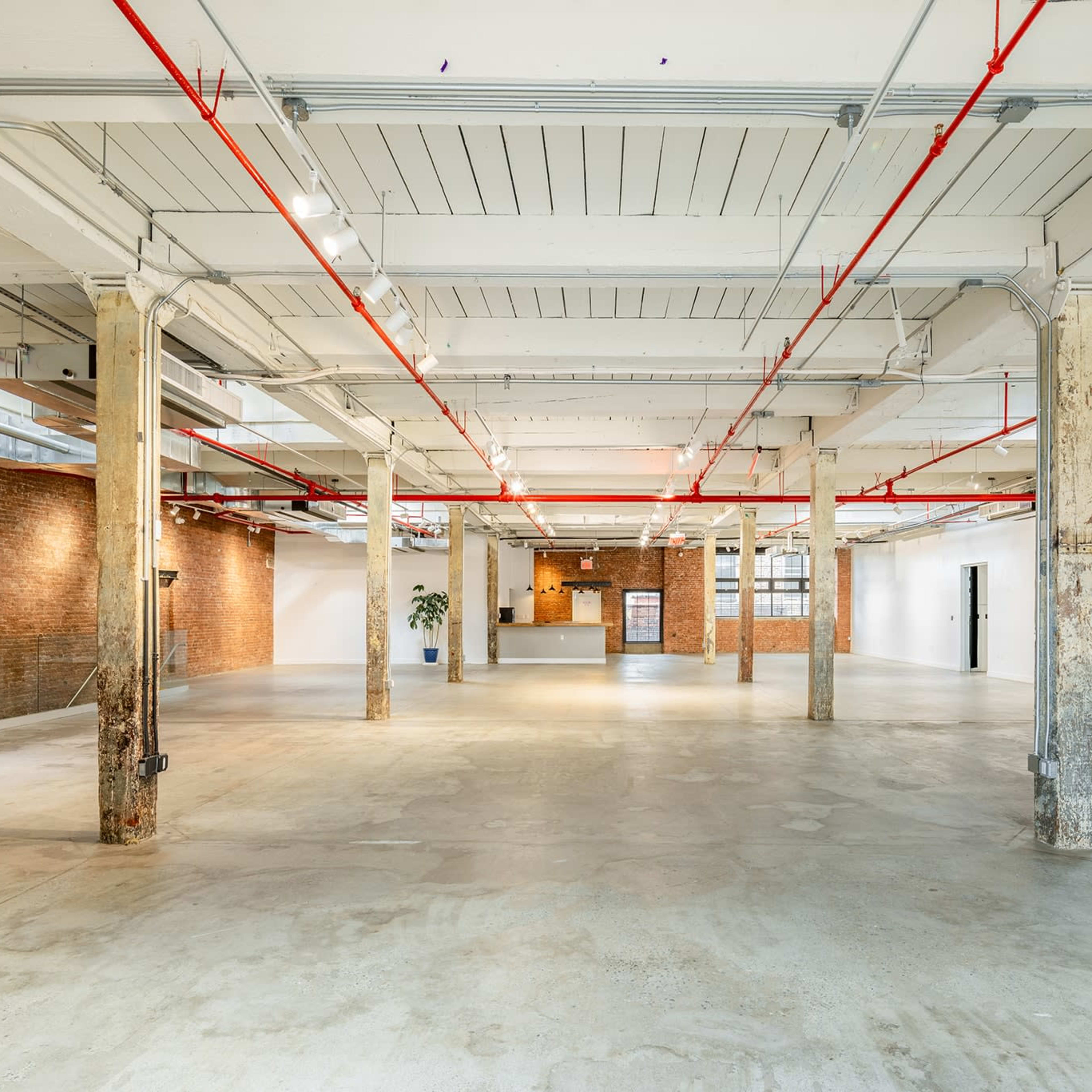 A spacious, unfinished industrial-style interior features exposed brick walls, concrete floors, and red ceiling pipes.