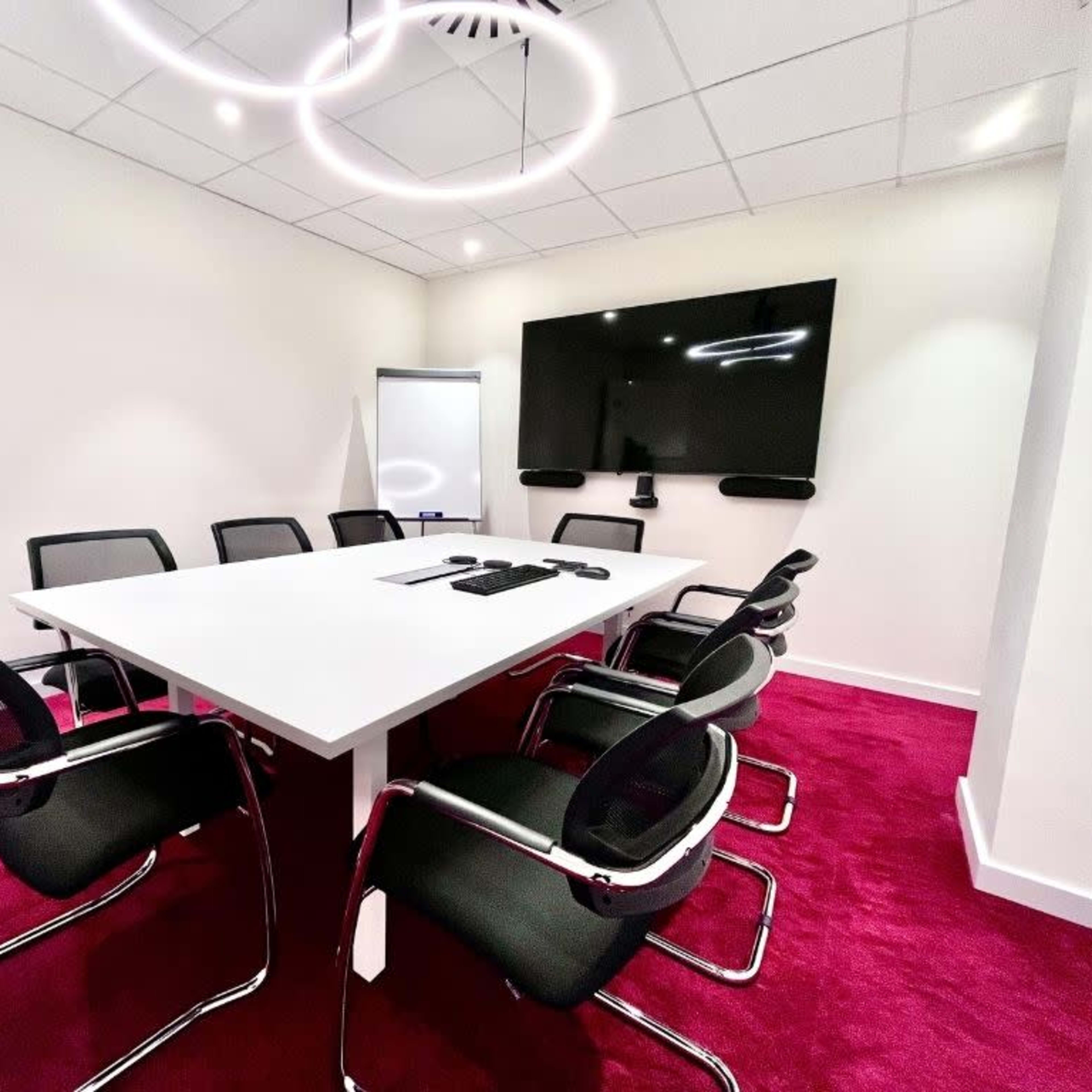 A modern conference room features a large white table surrounded by black chairs, with a wall-mounted screen and a flip chart in the background.