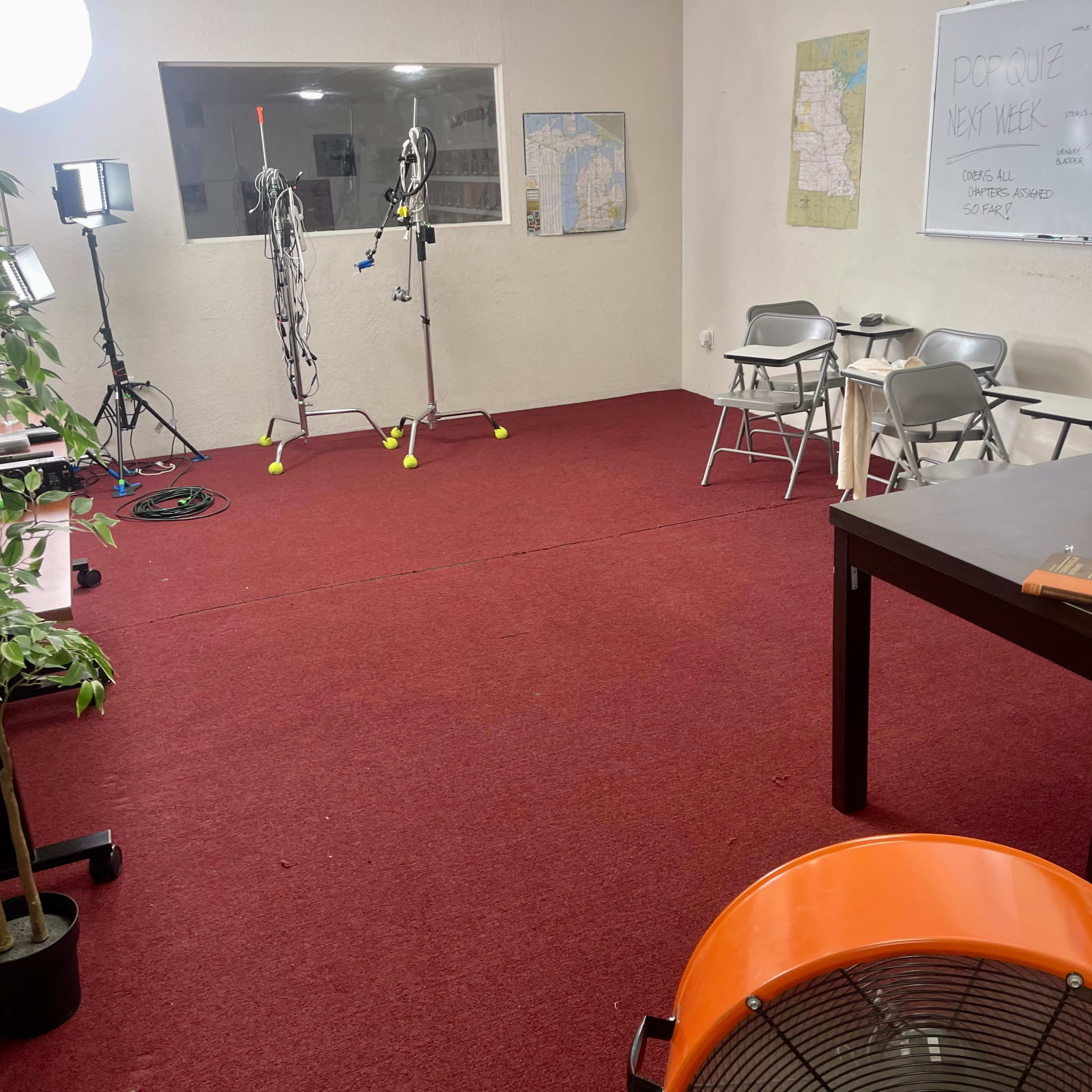 A sparsely furnished room with red carpet, equipped with filming equipment, a whiteboard, chairs, a table, a plant, and a fan.