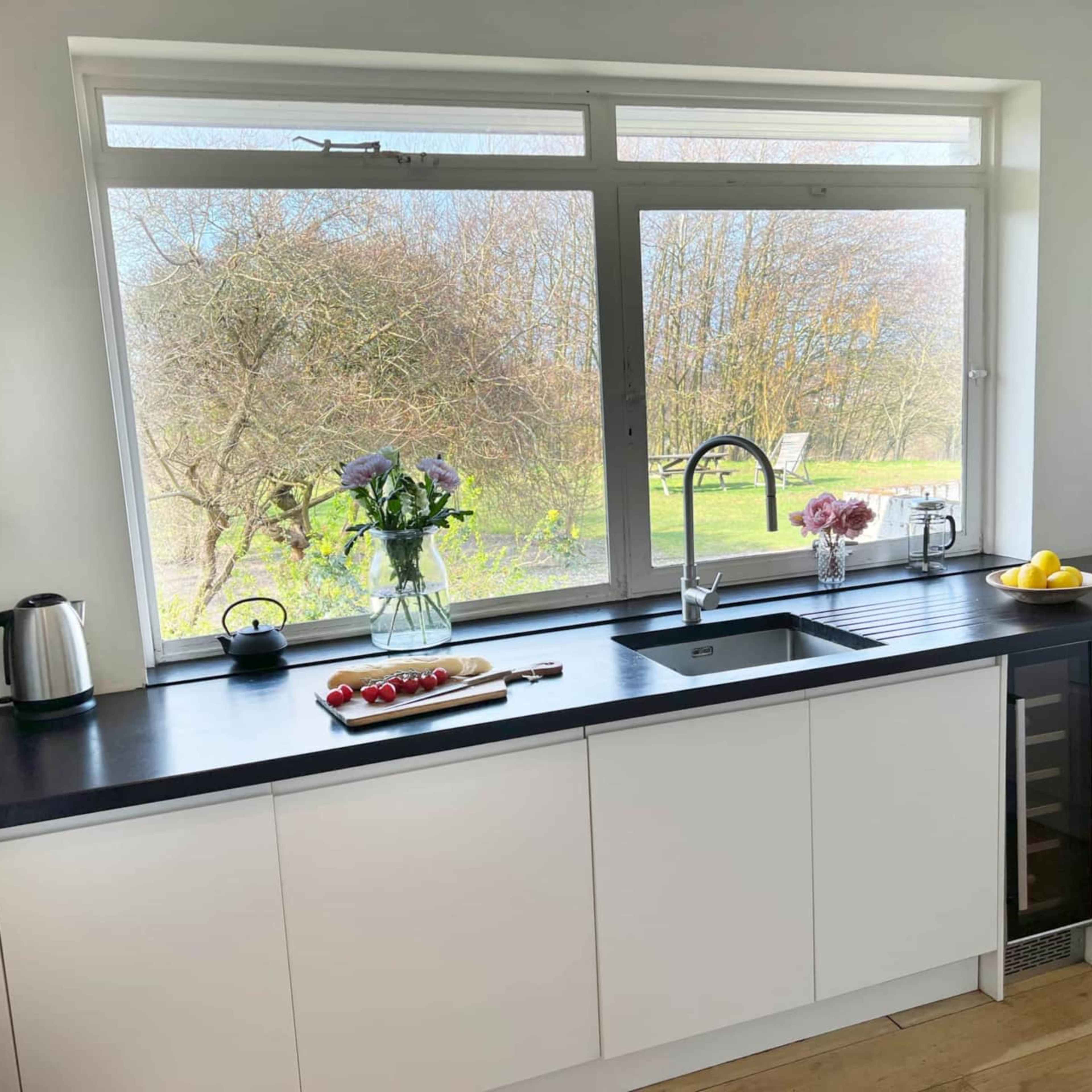 A modern kitchen features a window overlooking a garden, with flowers and fruit displayed on the counter.