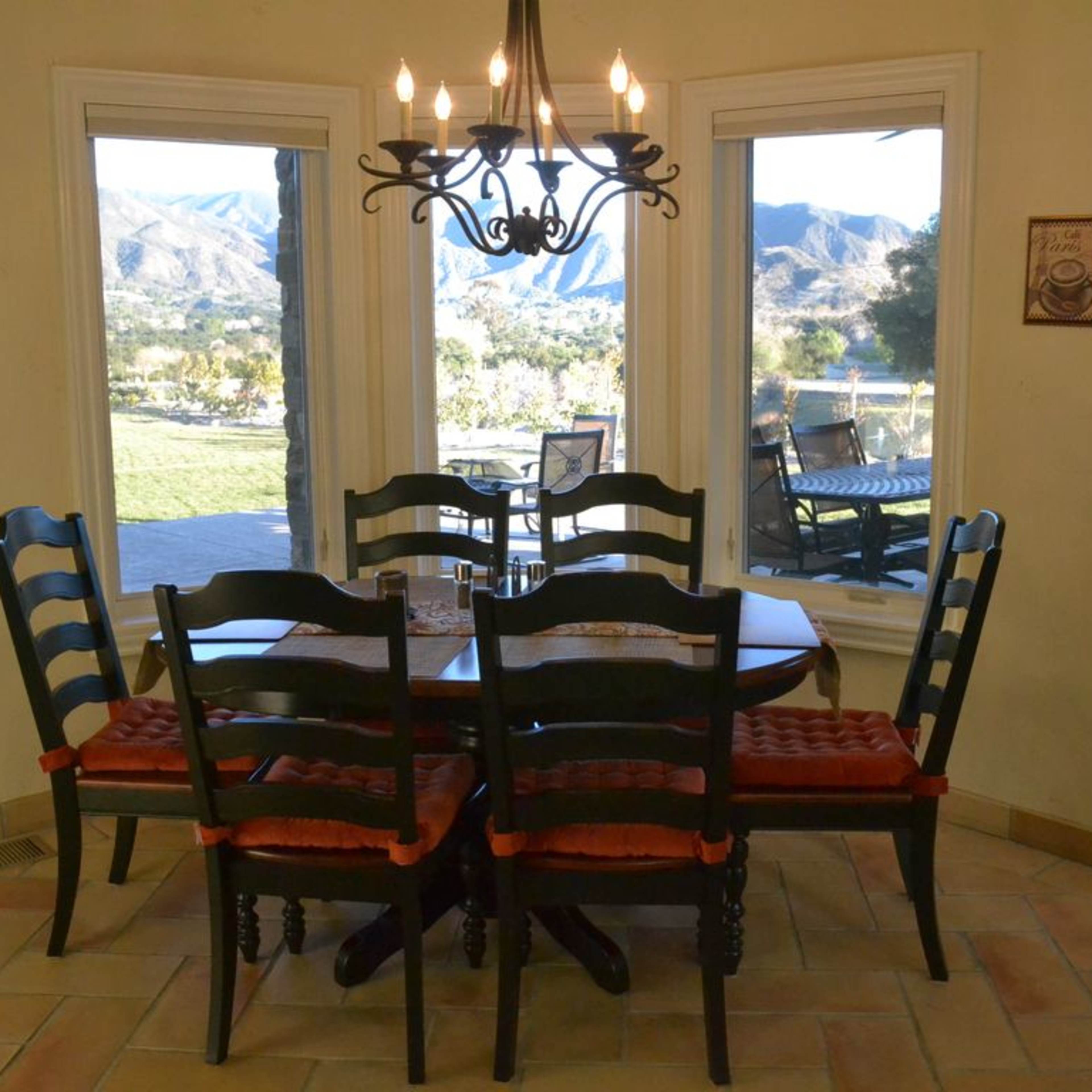 A dining table with six black chairs and red cushions is positioned near large windows offering a view of rolling hills and a patio.