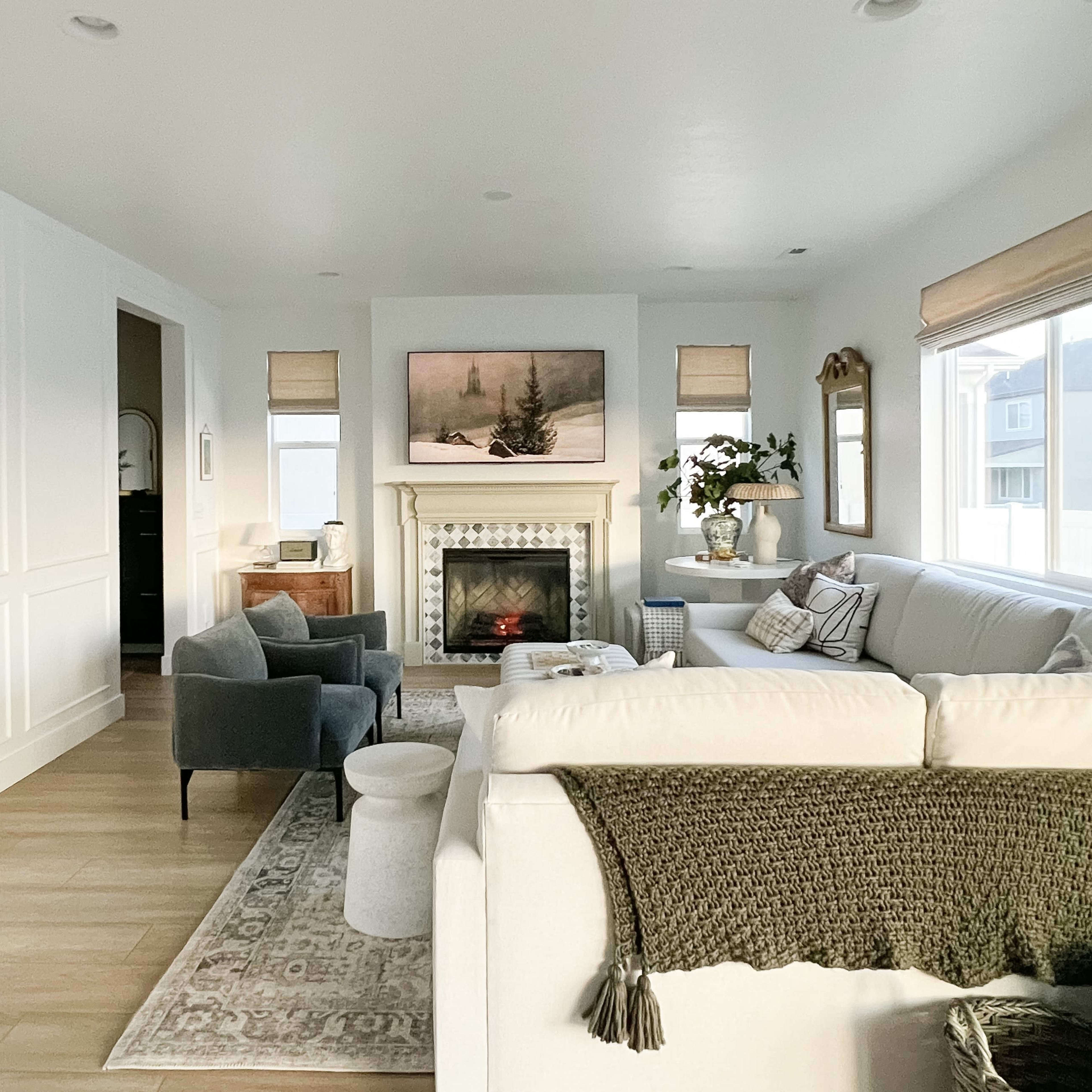 A bright living room featuring a large white sectional sofa, a fireplace with artwork above it, and two gray armchairs, all complemented by natural light from large windows.