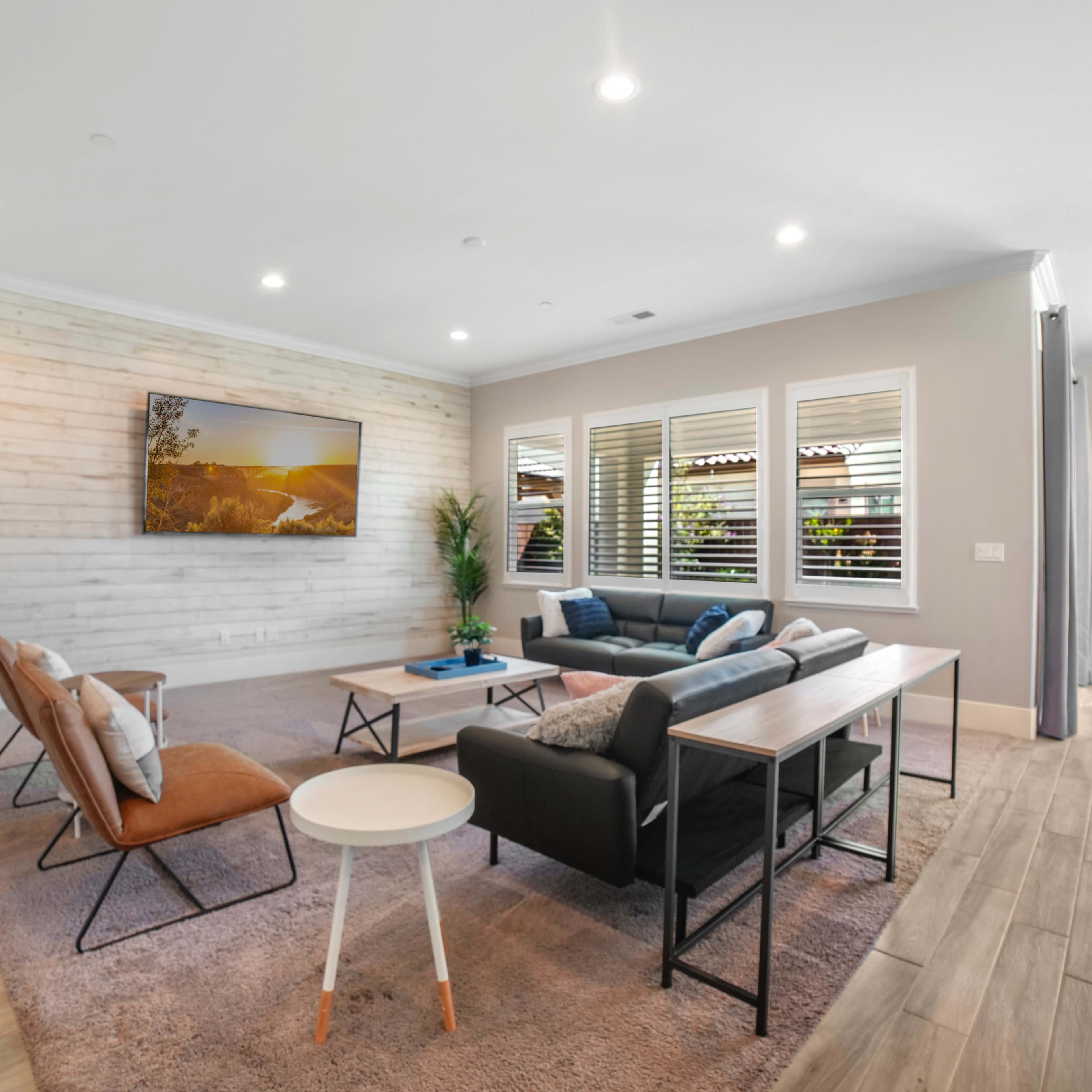 A modern living room featuring a combination of seating options, a wooden accent wall, and large windows with blinds.