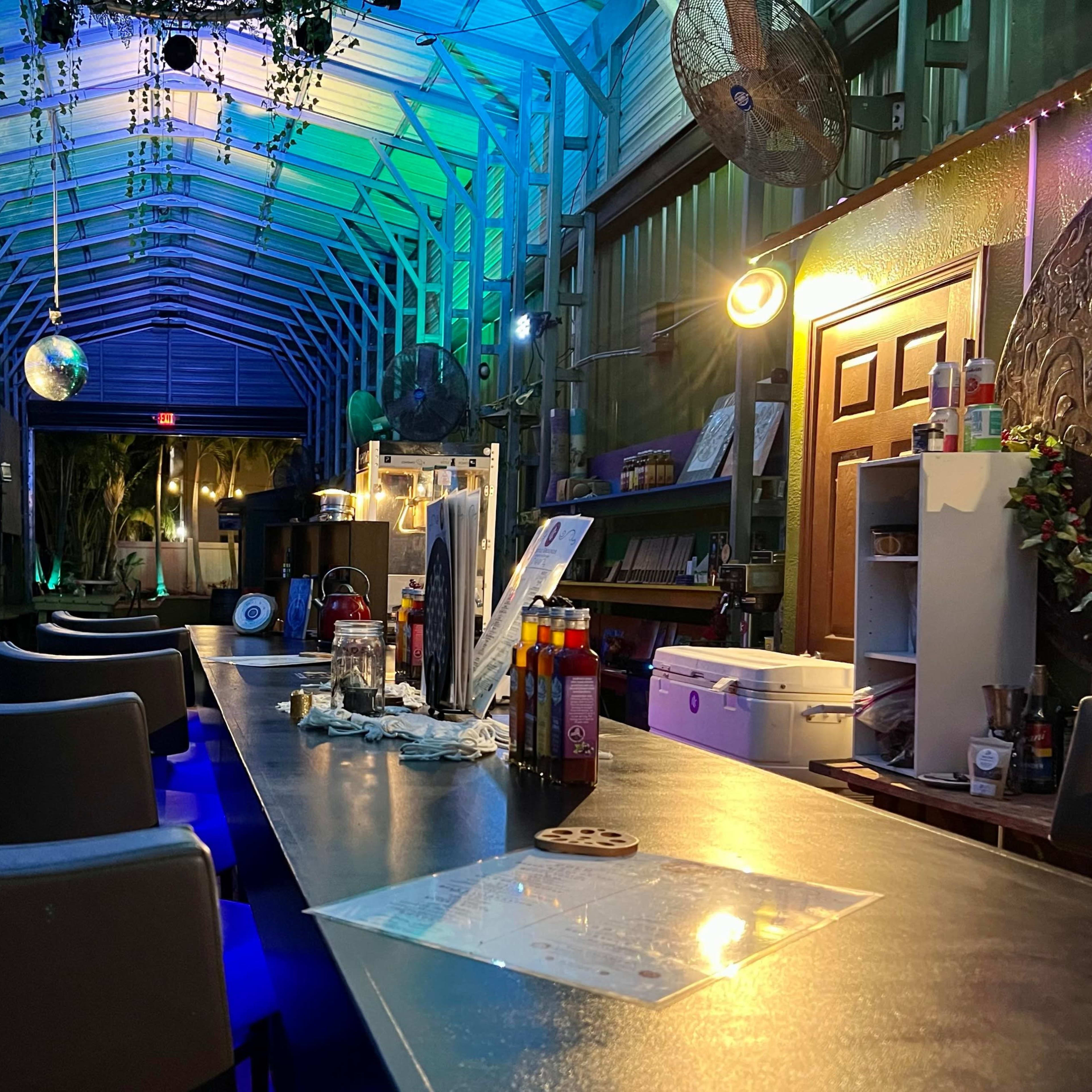 A bar area in a green, well-lit room with high ceilings, featuring a long counter, stools, and decorative lights.