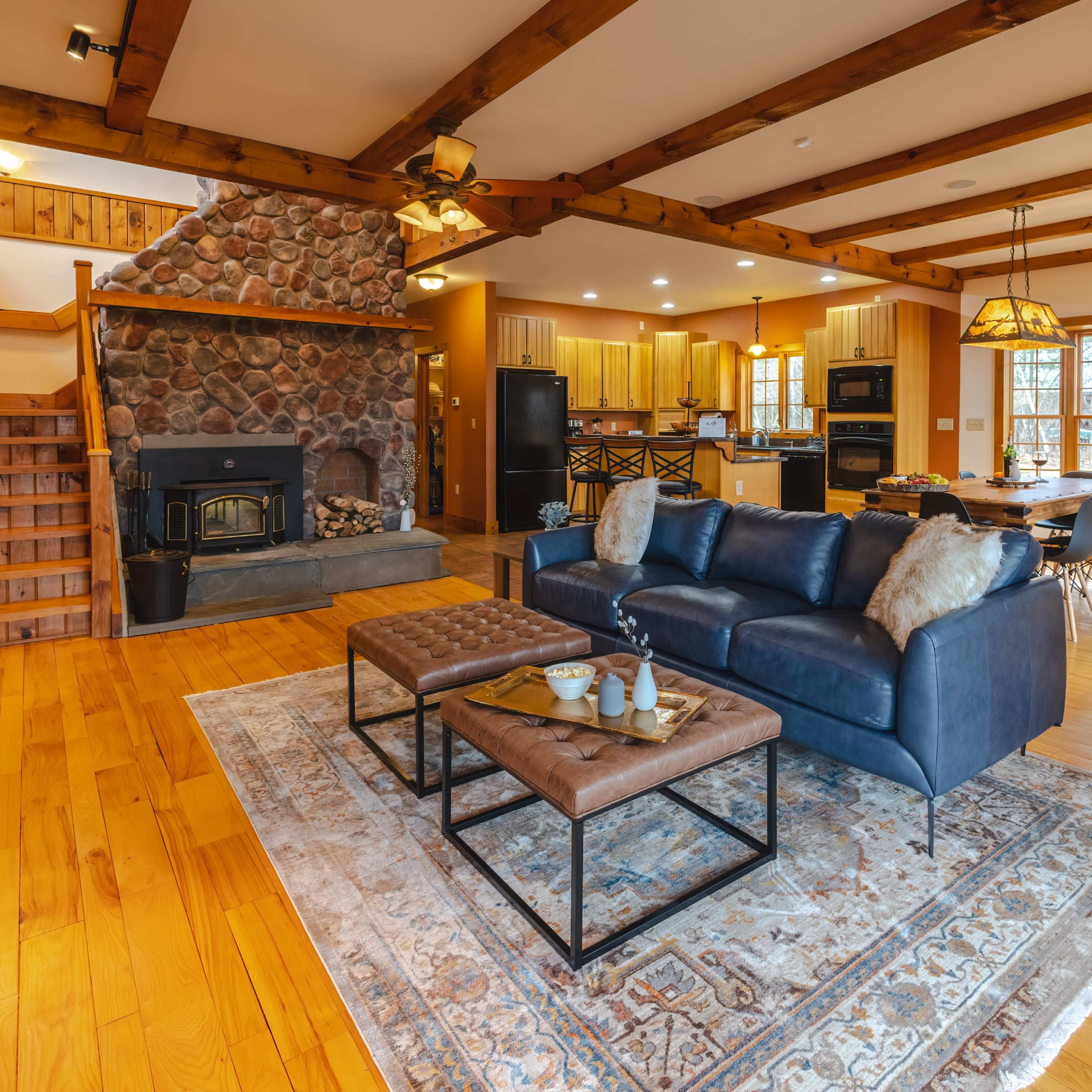 A spacious living area with a stone fireplace, wooden beams overhead, a leather sofa, and a dining area in the background.