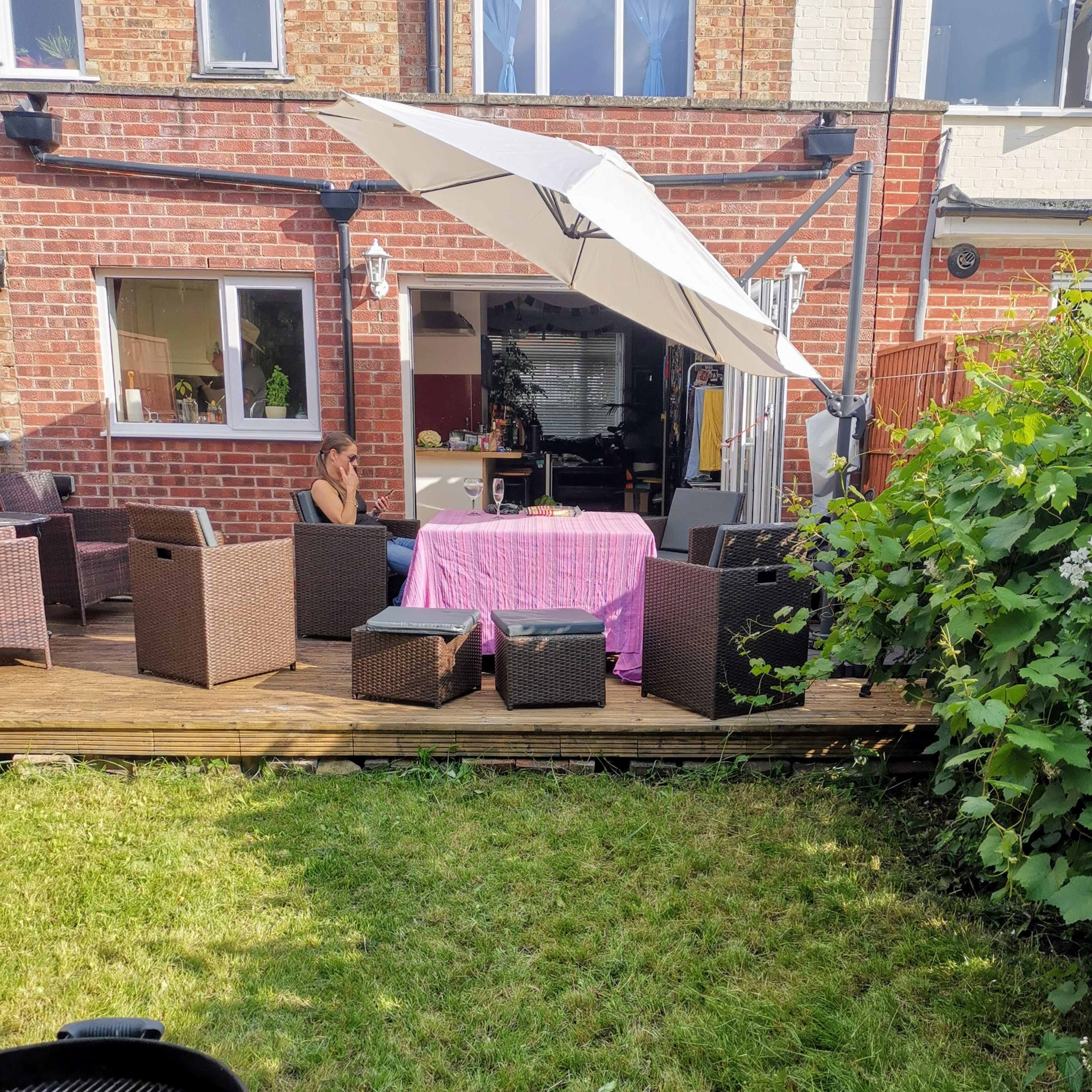 A backyard with a wooden deck furnished with dark wicker chairs and a table covered by a pink cloth, alongside a grill and surrounded by grass and greenery.