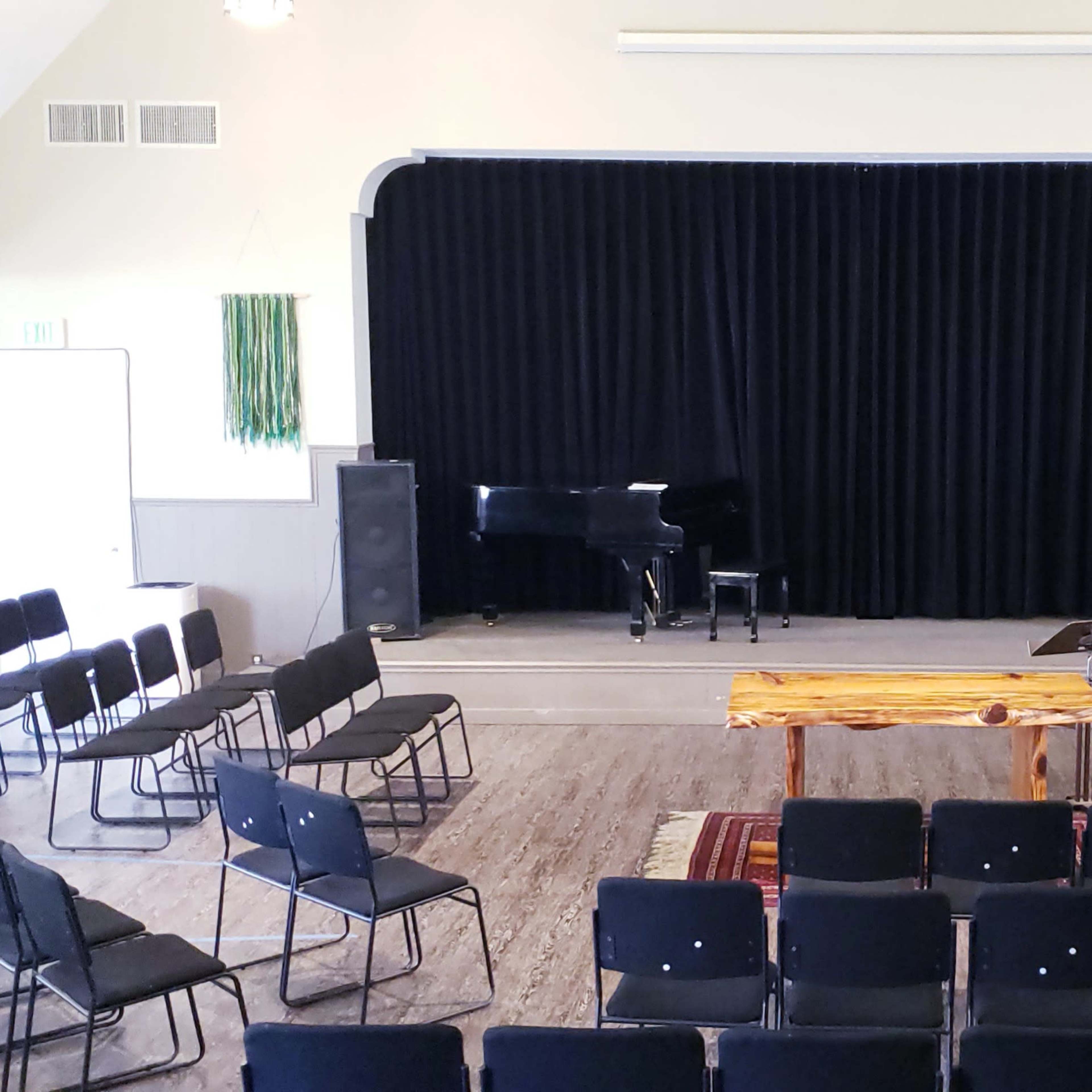 A performance space features rows of black folding chairs facing a wooden table and a grand piano, framed by black curtains and flanked by speakers.