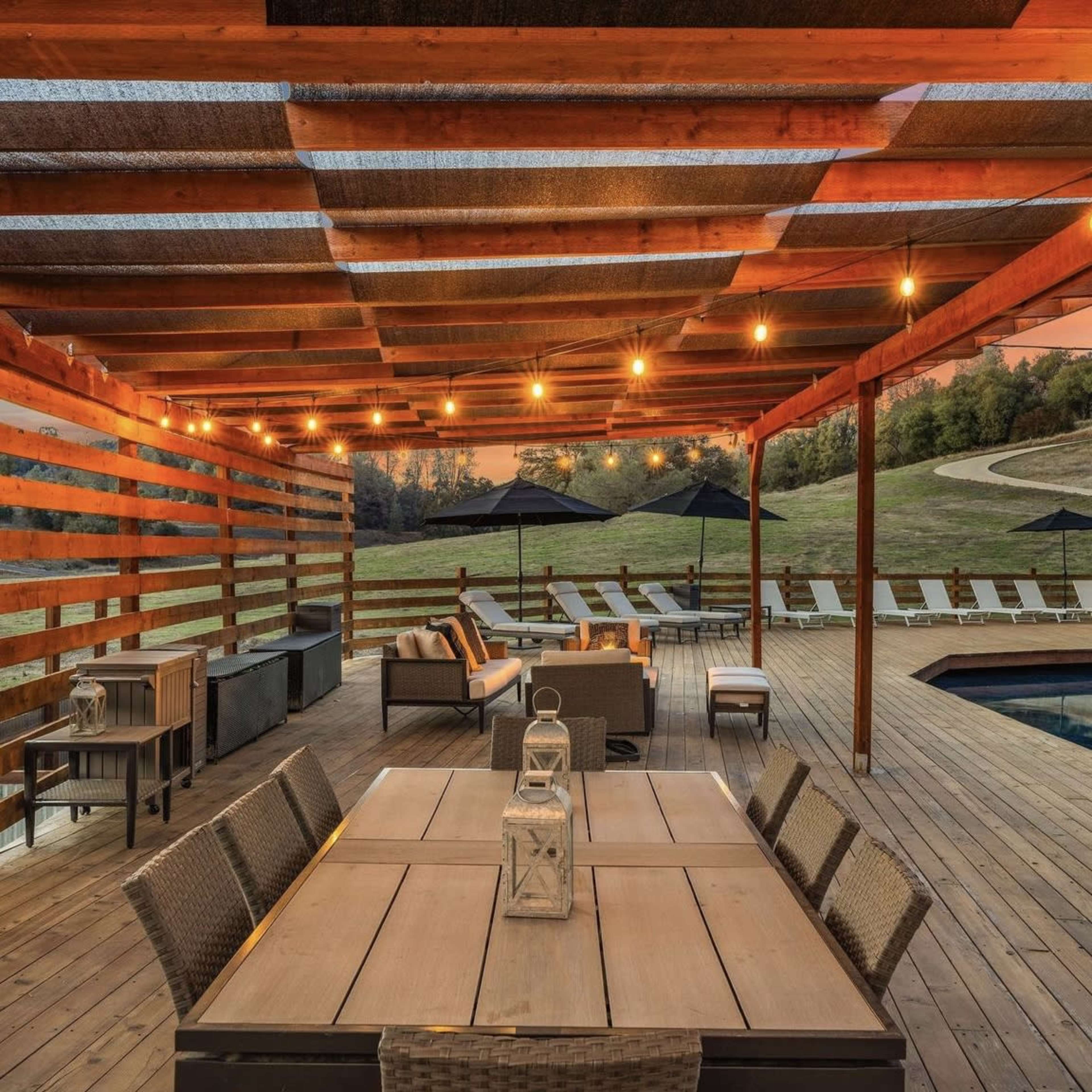 A wooden patio area with seating and a dining table, illuminated by string lights, overlooking a pool and lounge chairs in a grassy landscape.