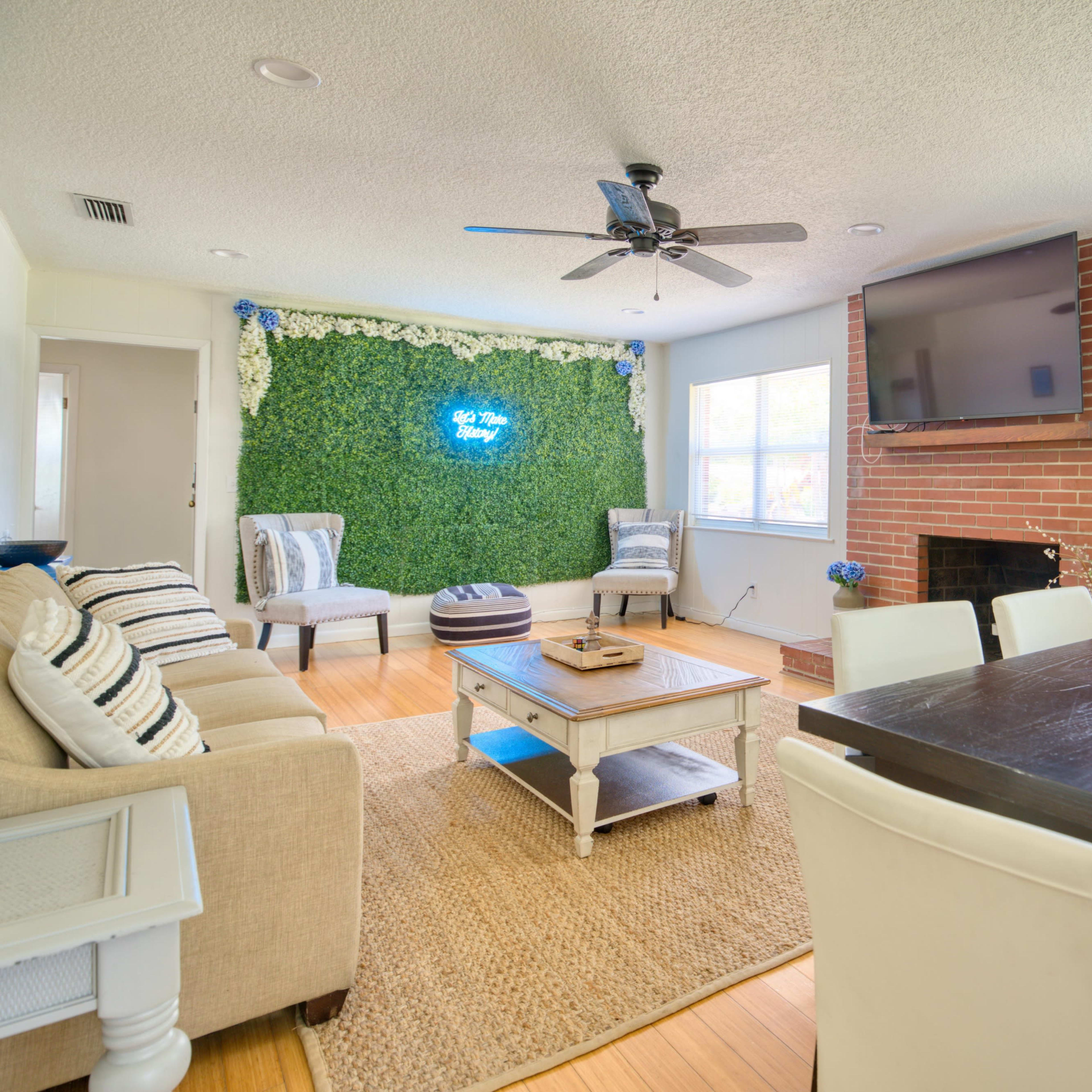 A living room with a green wall featuring decorative plants, a brick fireplace, a television, a sofa, and a dining table with chairs.