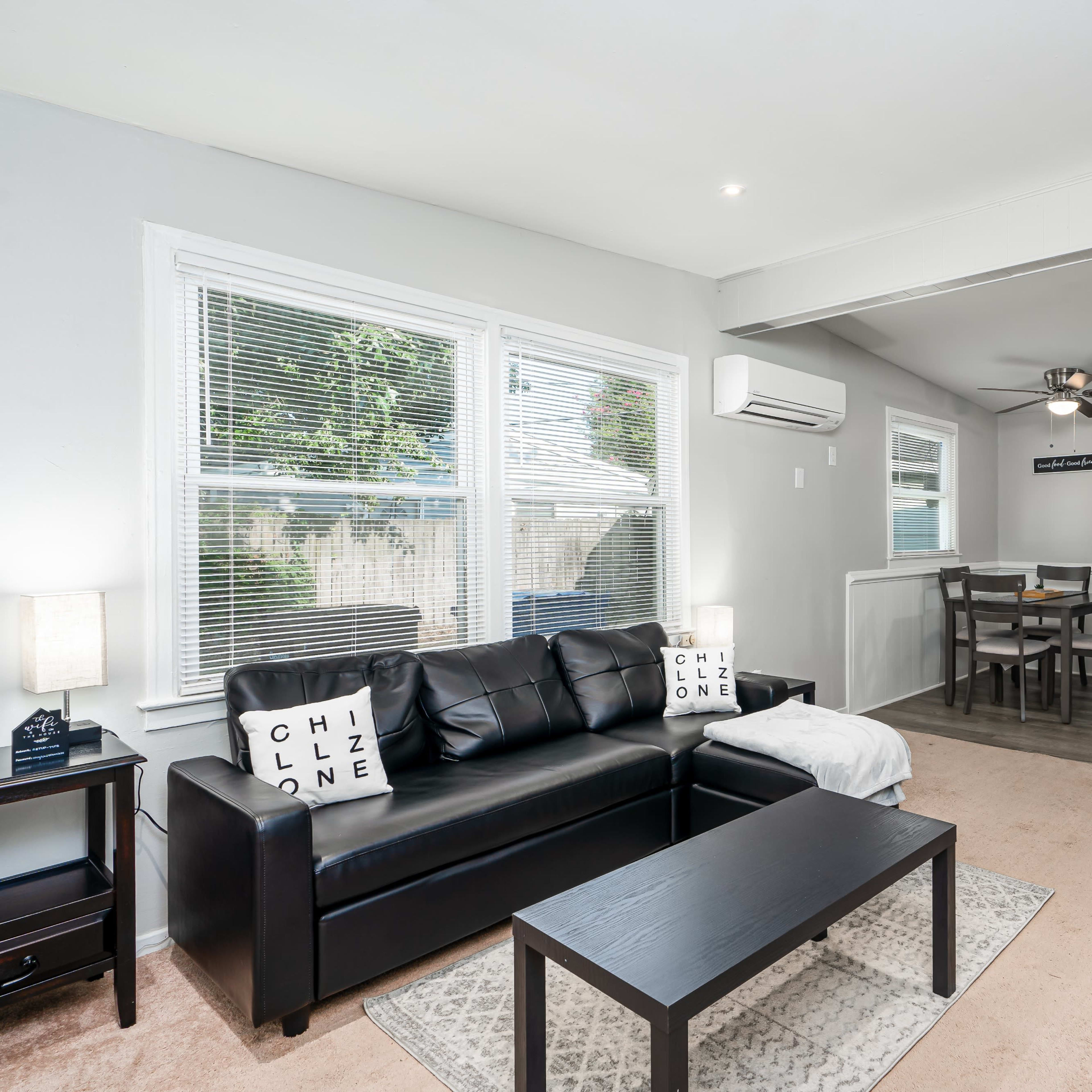 A modern living room features a black leather couch, a coffee table, and a dining area with a circular mirror on the wall.