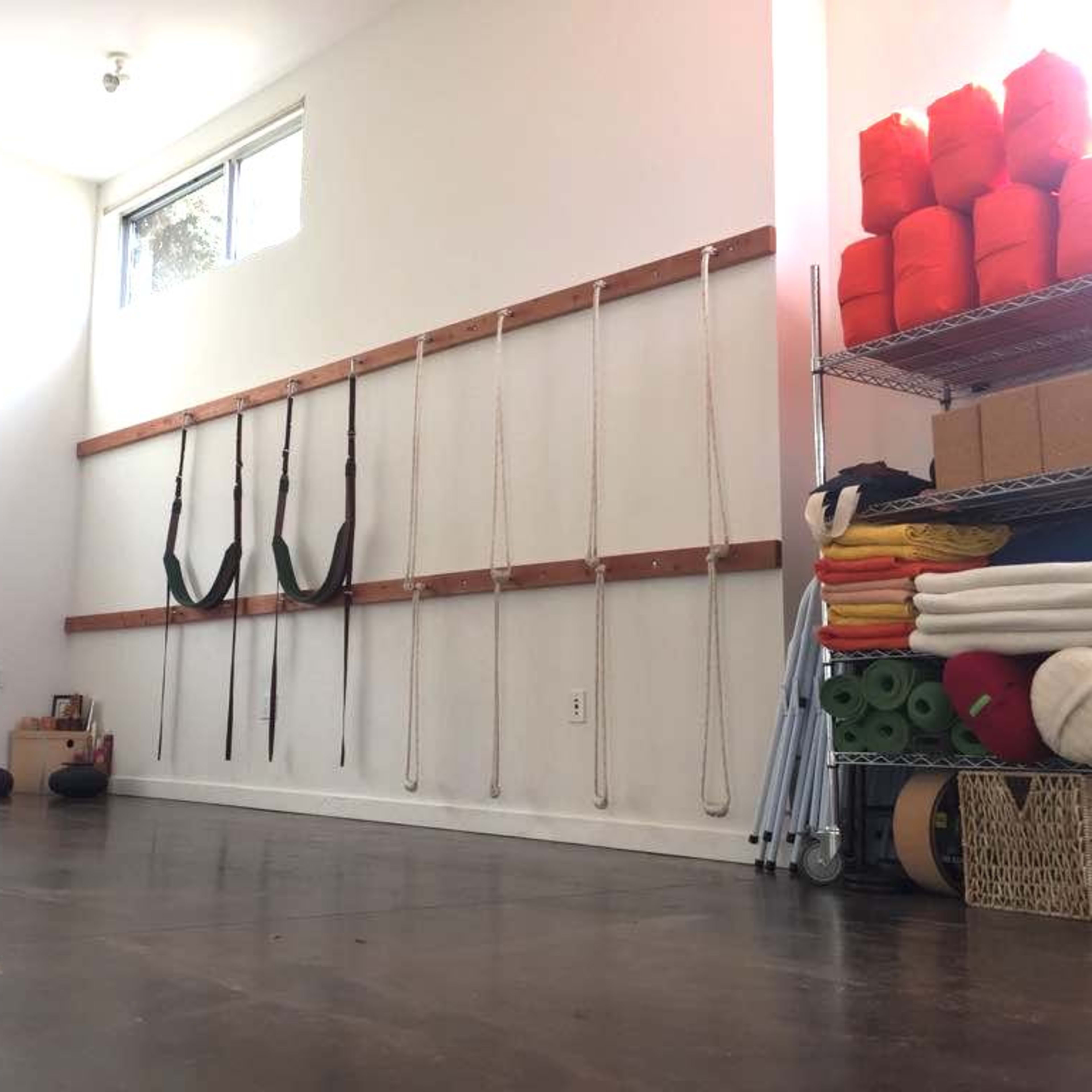 A bright, minimalist room with a wooden wall-mounted system for yoga straps and ropes, alongside neatly organized exercise mats and props stored on shelves.