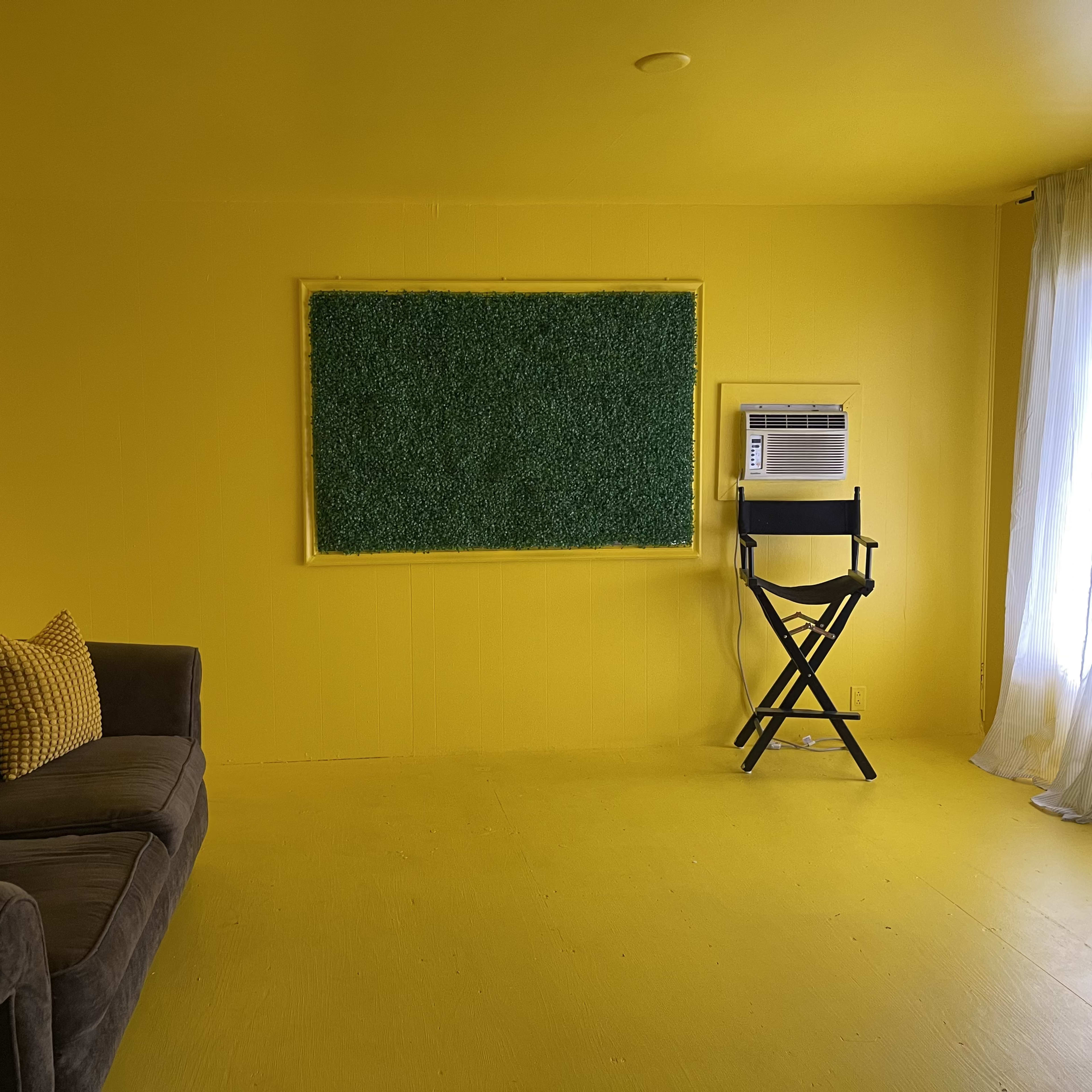 The room features yellow walls and flooring, a gray sofa with yellow pillows, a black director's chair, and a green framed board on one wall.